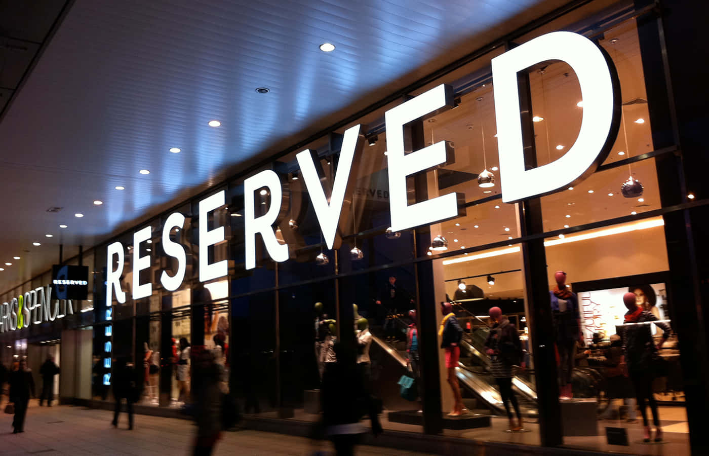 Reserved 桌布 壁紙 — 夜晚明亮照明的RESERVED商店，玻璃窗反射霓虹燈光和模糊的行人走過。