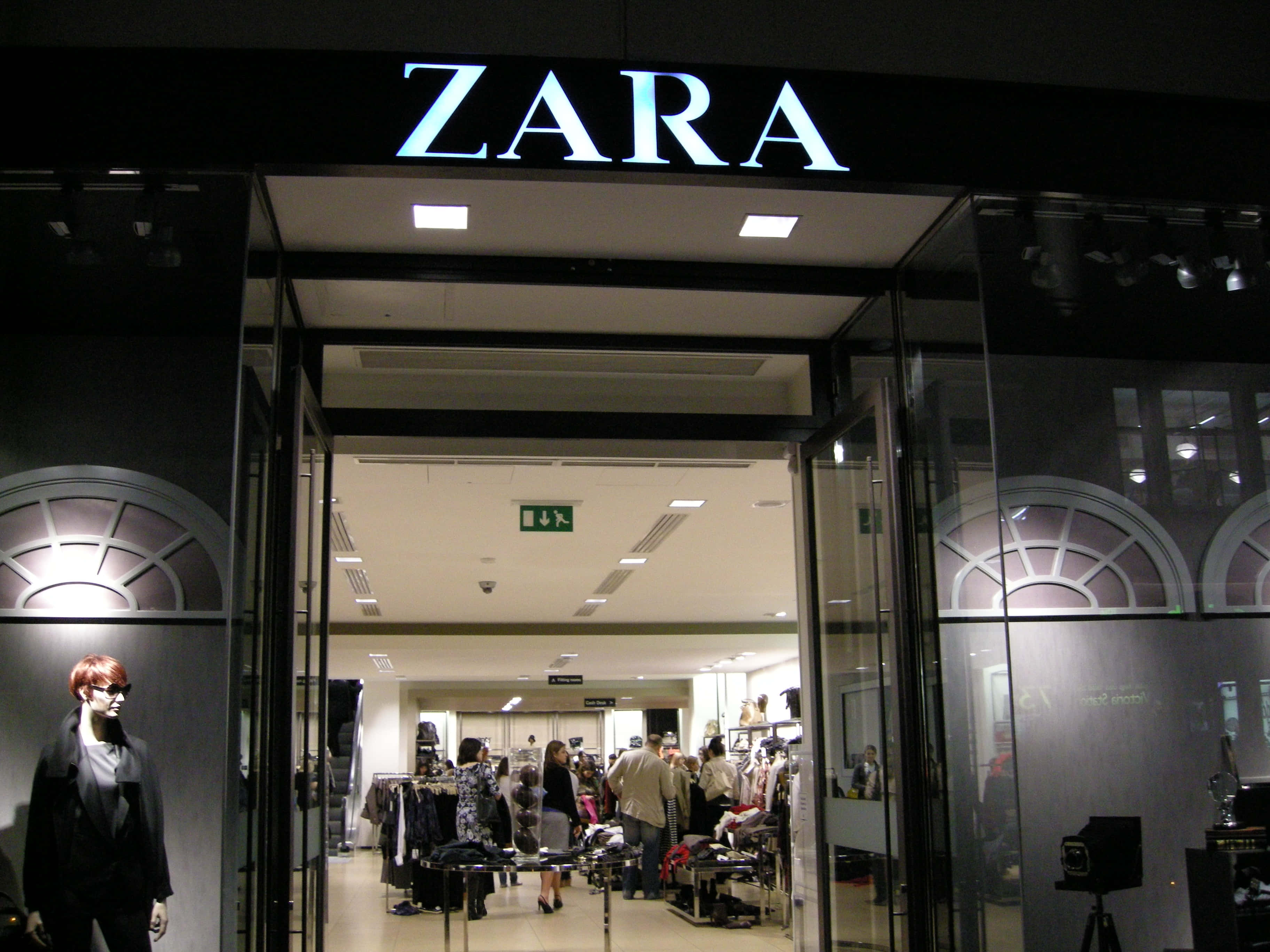 Zara 桌布 壁紙 — 夜晚明亮照明的Zara店鋪入口，玻璃門敞開，顧客在店內瀏覽貨架。