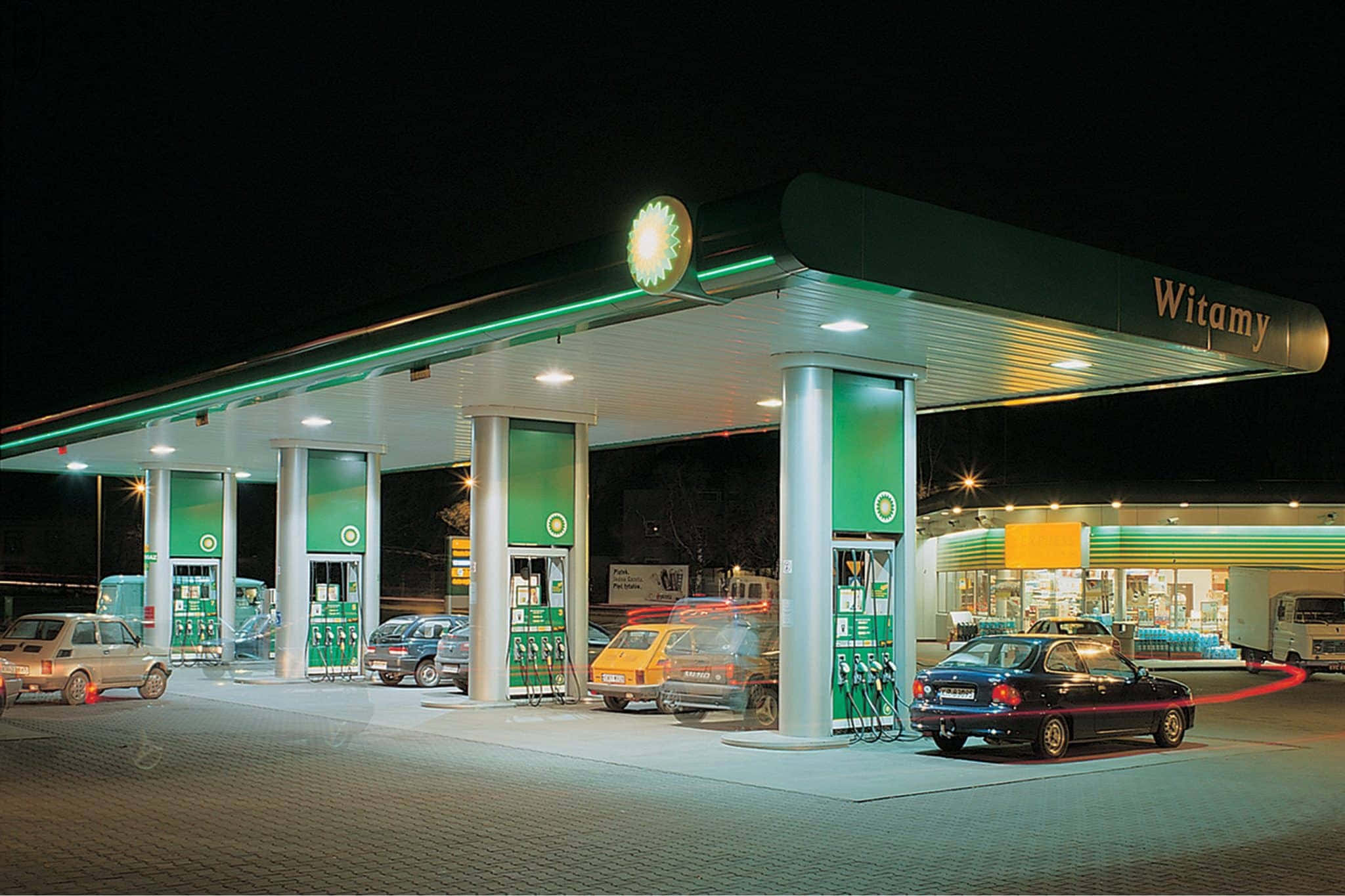 Petrol Station 桌布 壁紙 — 夜晚明亮照明的加油站，多輛汽車在發光的綠色標誌和聚光燈下加油。