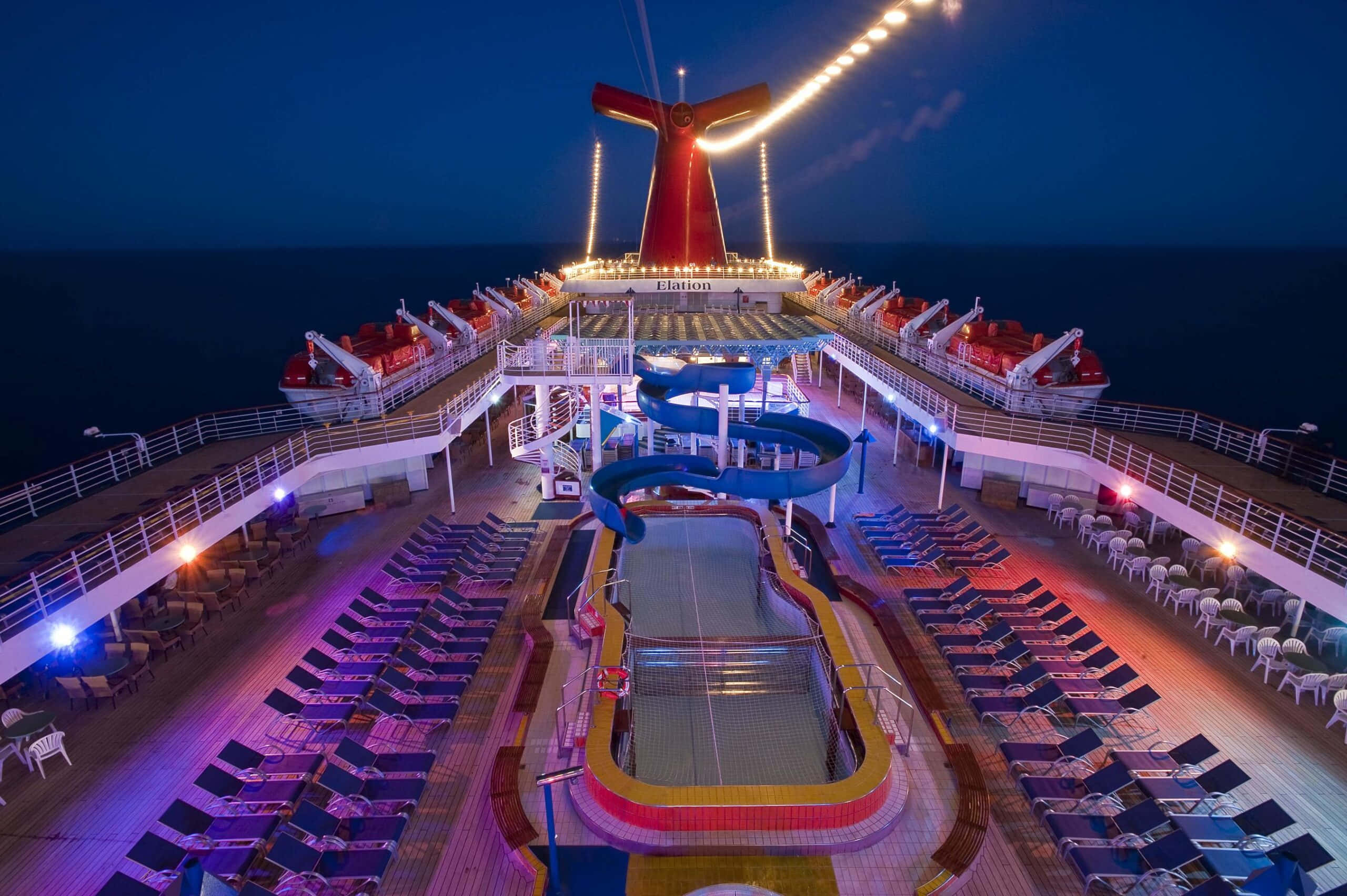 Carnival Conquest 桌布 壁紙 — 夜晚明亮照明的郵輪甲板上，中央有一個帶藍色水上滑梯的游泳池，周圍排列著躺椅，上方是漆黑的天空。