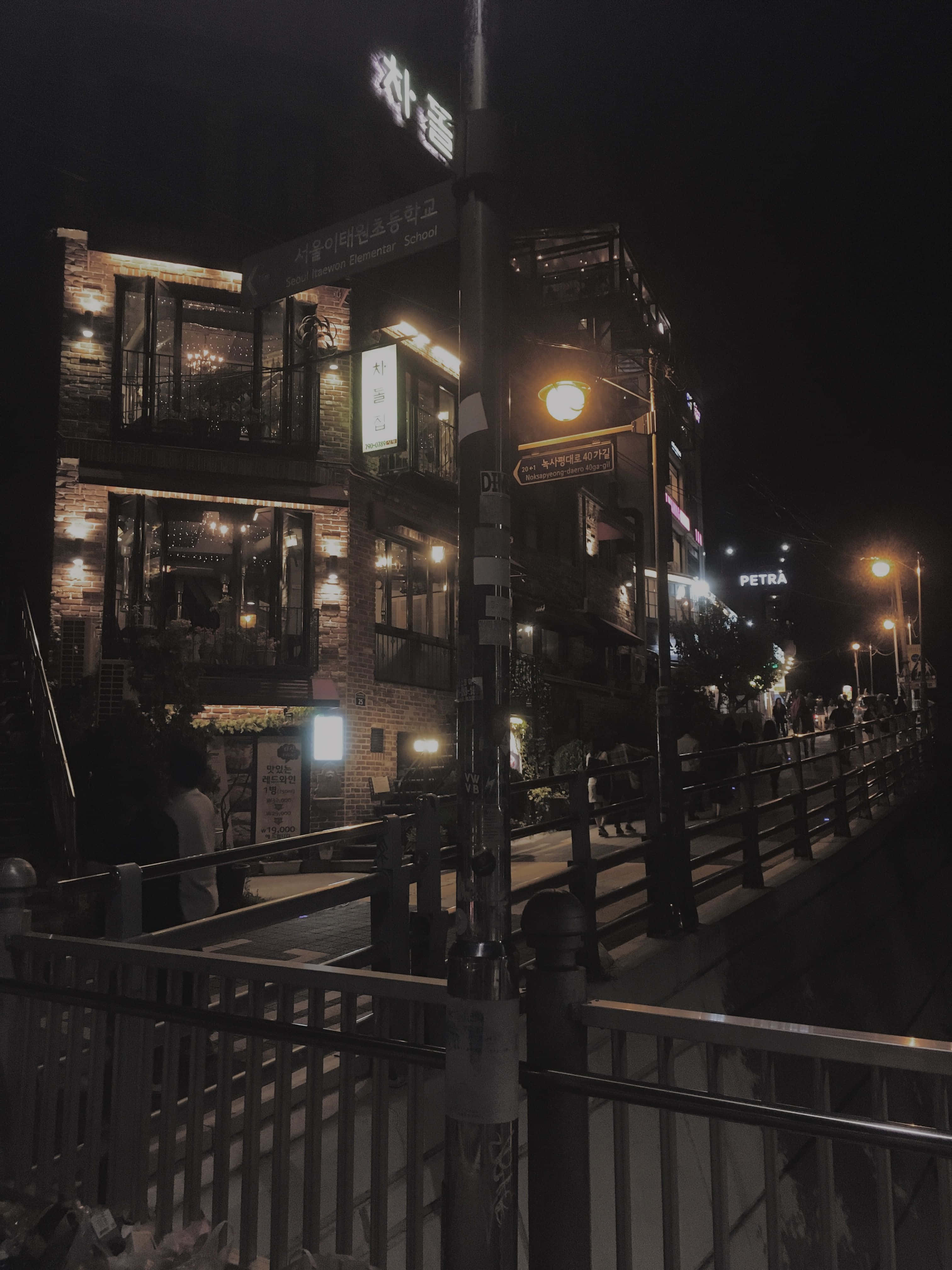 暗黑風格 桌布 壁紙 — 夜晚昏暗的城市街道，發光的霓虹燈招牌和溫暖的路燈照亮磚造建築和行人。