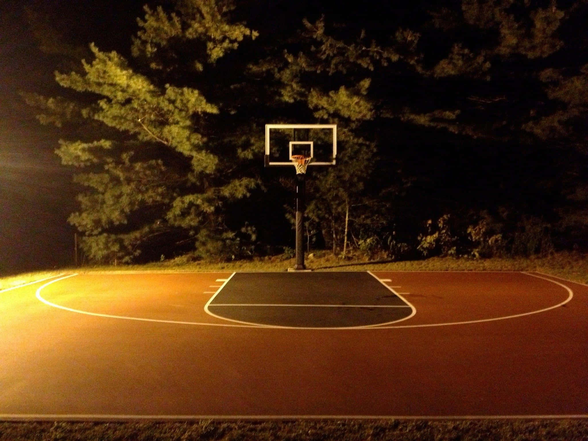 Basketball Court 桌布 壁紙 — 夜晚昏暗的戶外籃球場，籃框位於聚光燈下中央，周圍環繞著黑暗的樹木。