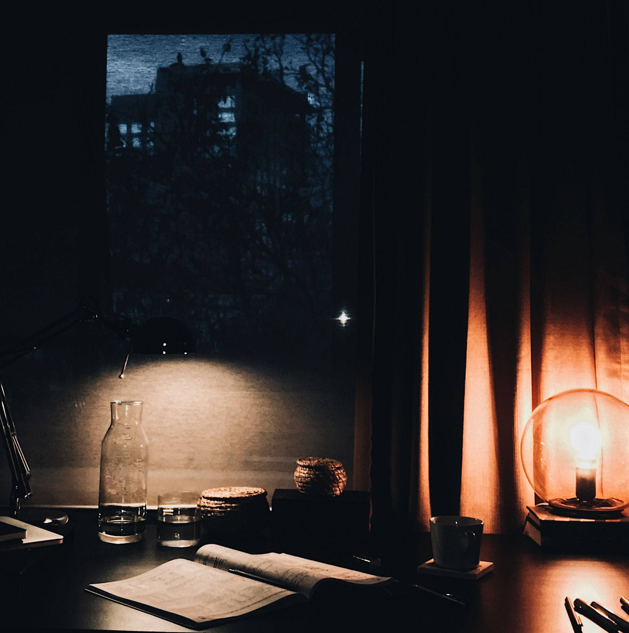 Desk 桌布 壁紙 — 夜晚昏暗的書桌，一盞發光的燈在黑暗的窗景前將溫暖的光線投射在打開的書本和玻璃水瓶上。