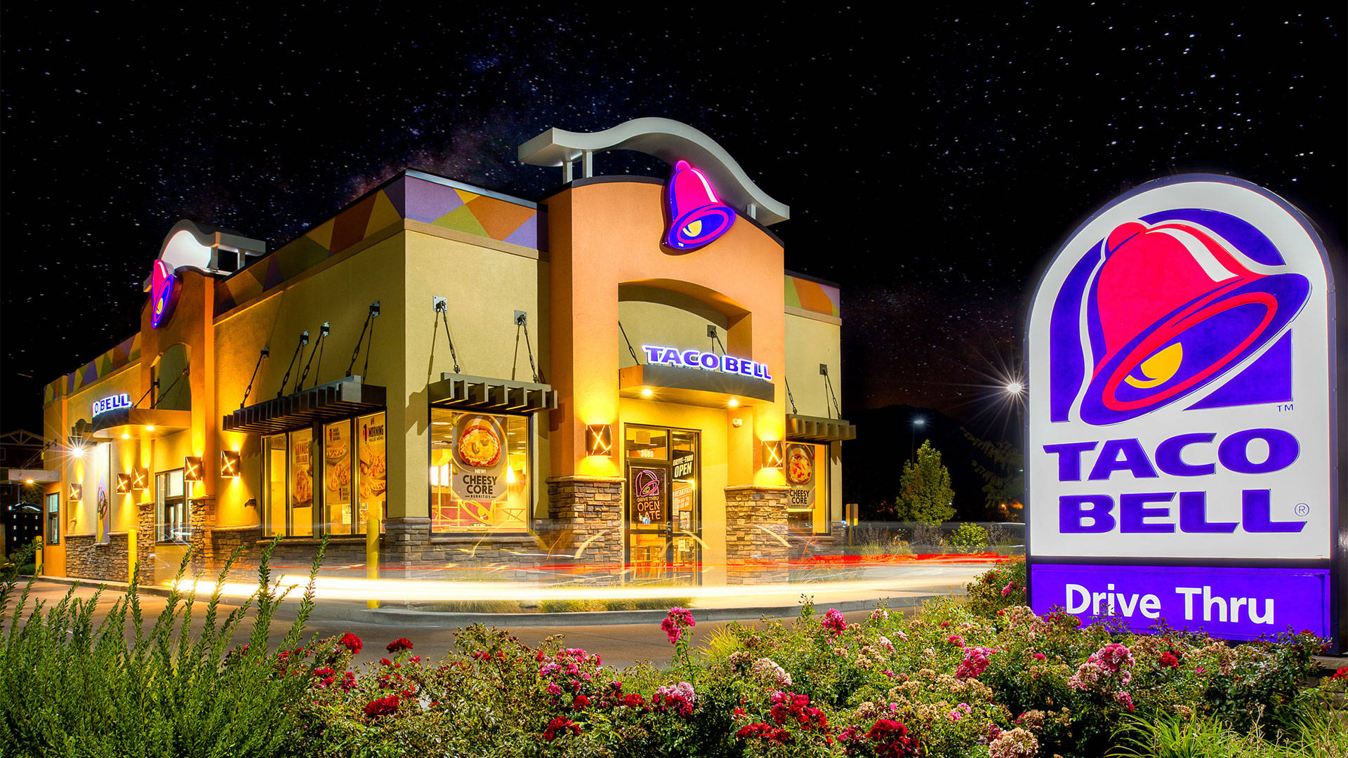 塔可鐘 桌布 壁紙 — 夜晚星空下，一間燈火通明的Taco Bell餐廳，發光的招牌和動態模糊的車輛駛過。