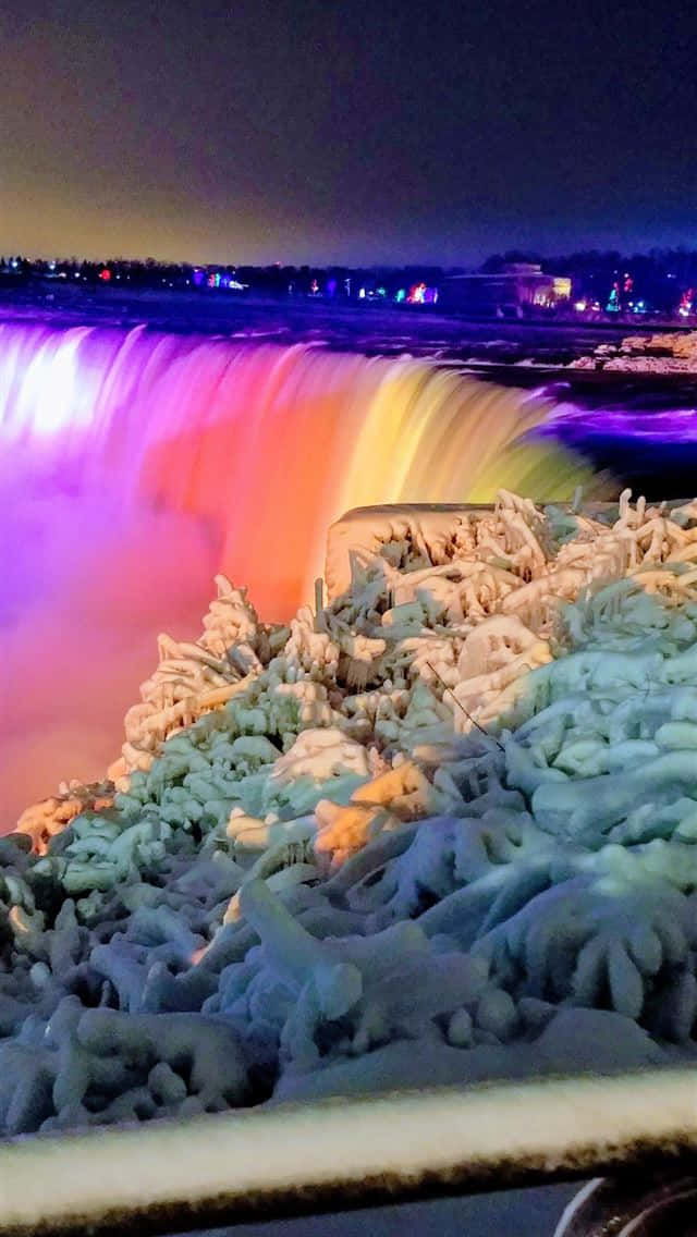 雪 桌布 壁紙 — 夜晚時分，一條結冰的瀑布被鮮豔的紫色、橙色和黃色燈光照亮，在冰層上投射出色彩繽紛的倒影。