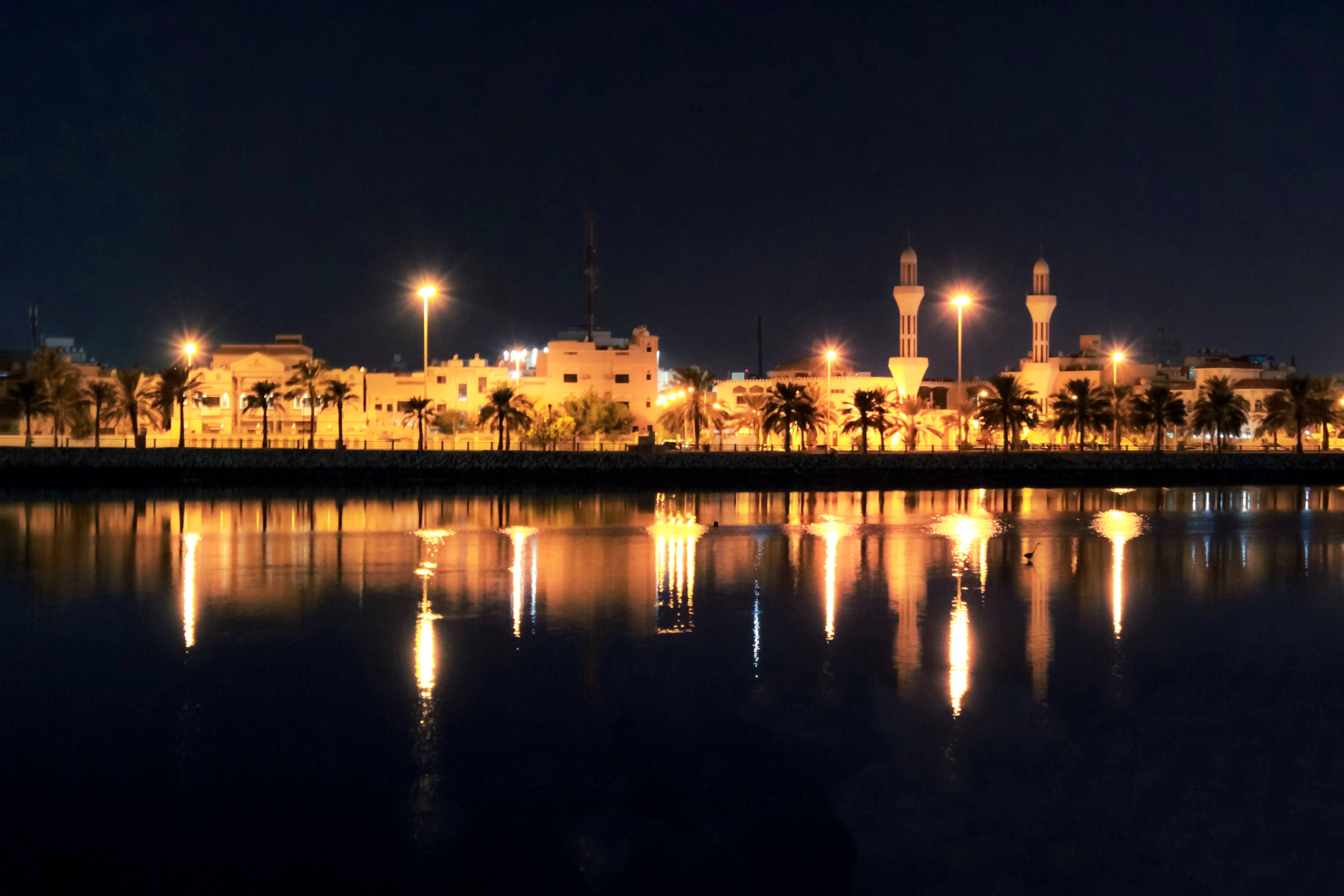 Bahrain 桌布 壁紙 — 夜晚景色，照亮嘅清真寺宣禮塔同棕櫚樹倒映喺平靜嘅水面，天空漆黑一片。