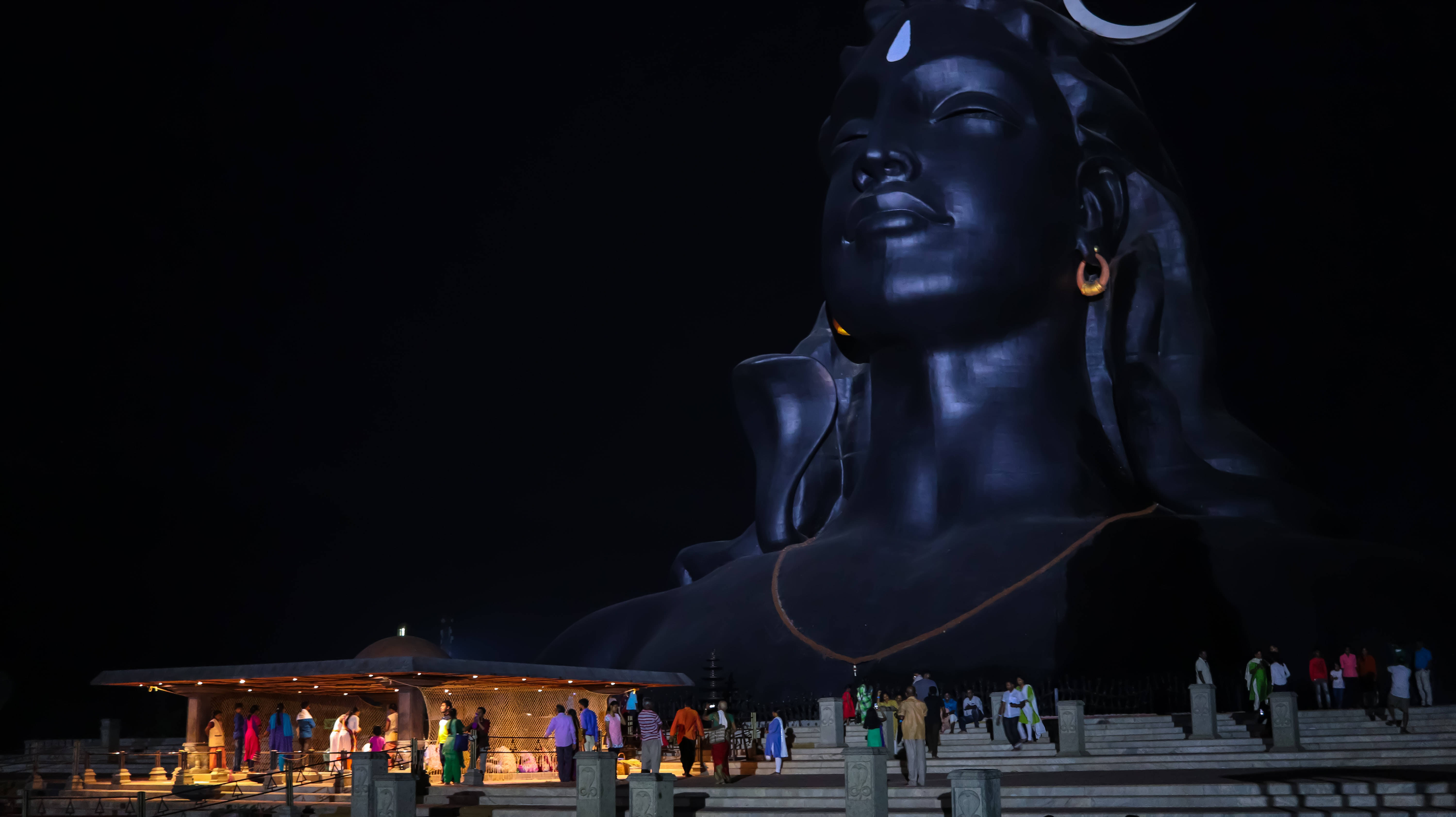 adiyogi shiva 桌布 壁紙 — 夜晚暗空下，巨大發光的濕婆神像，下方階梯上有遊客。