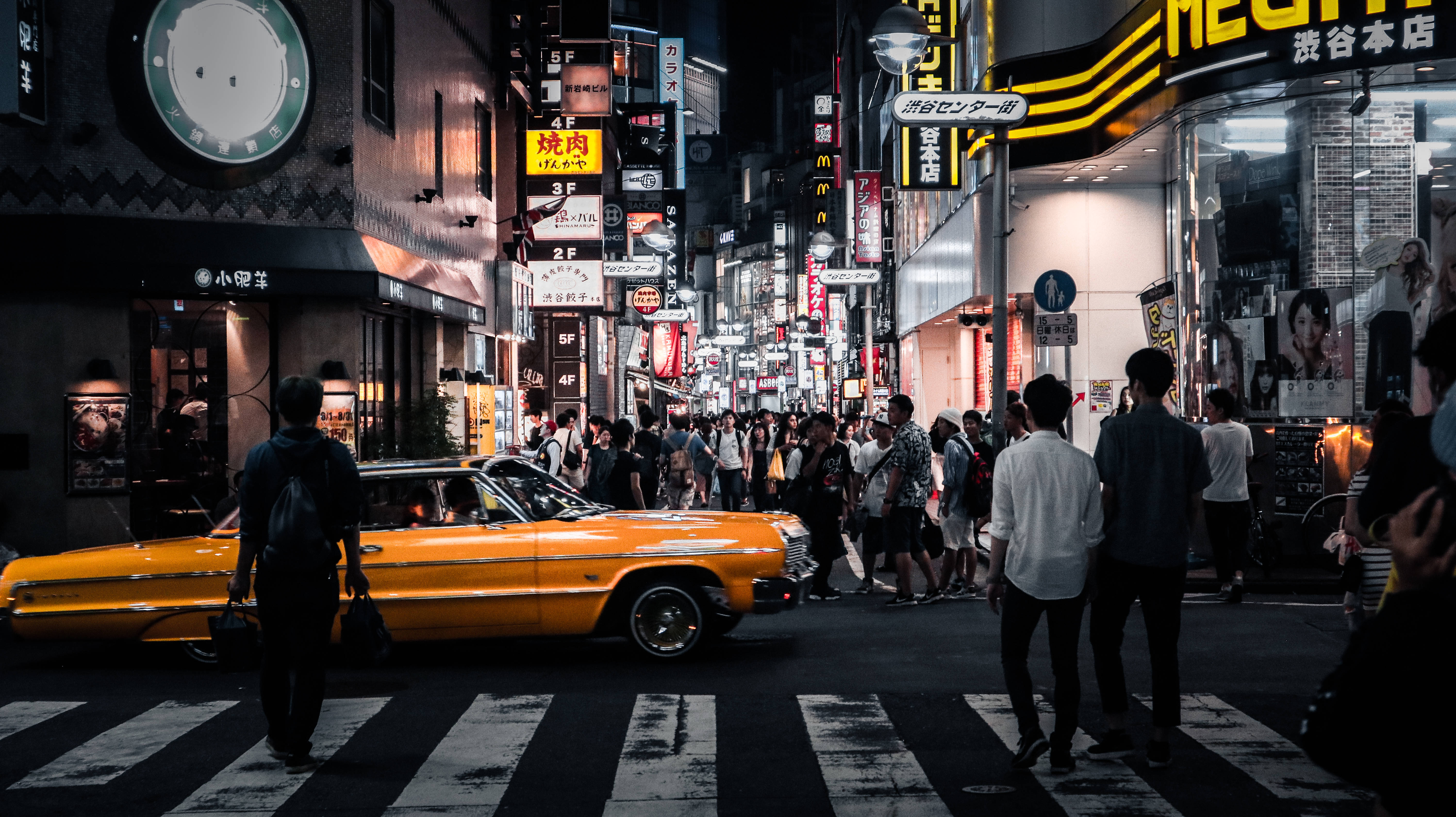 Lowrider 桌布 壁紙 — 夜晚東京一條充滿活力的街道，一輛亮黃色的經典汽車在閃爍的霓虹燈招牌和人群中穿越行人穿越道。