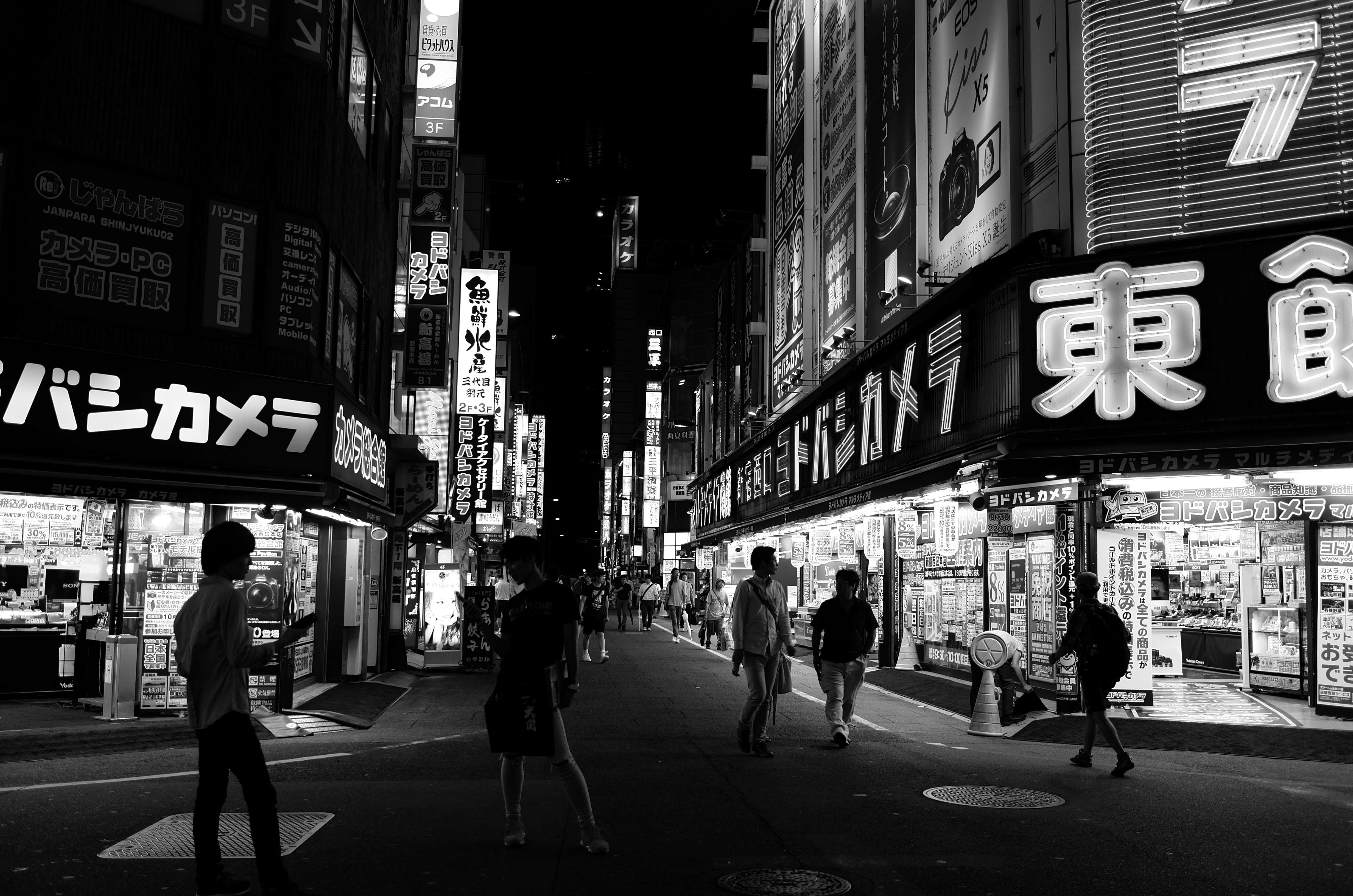 日本黑白 桌布 壁紙 — 夜晚東京一條繁忙的霓虹燈街道，行人走過發光的商店櫥窗和招牌。
