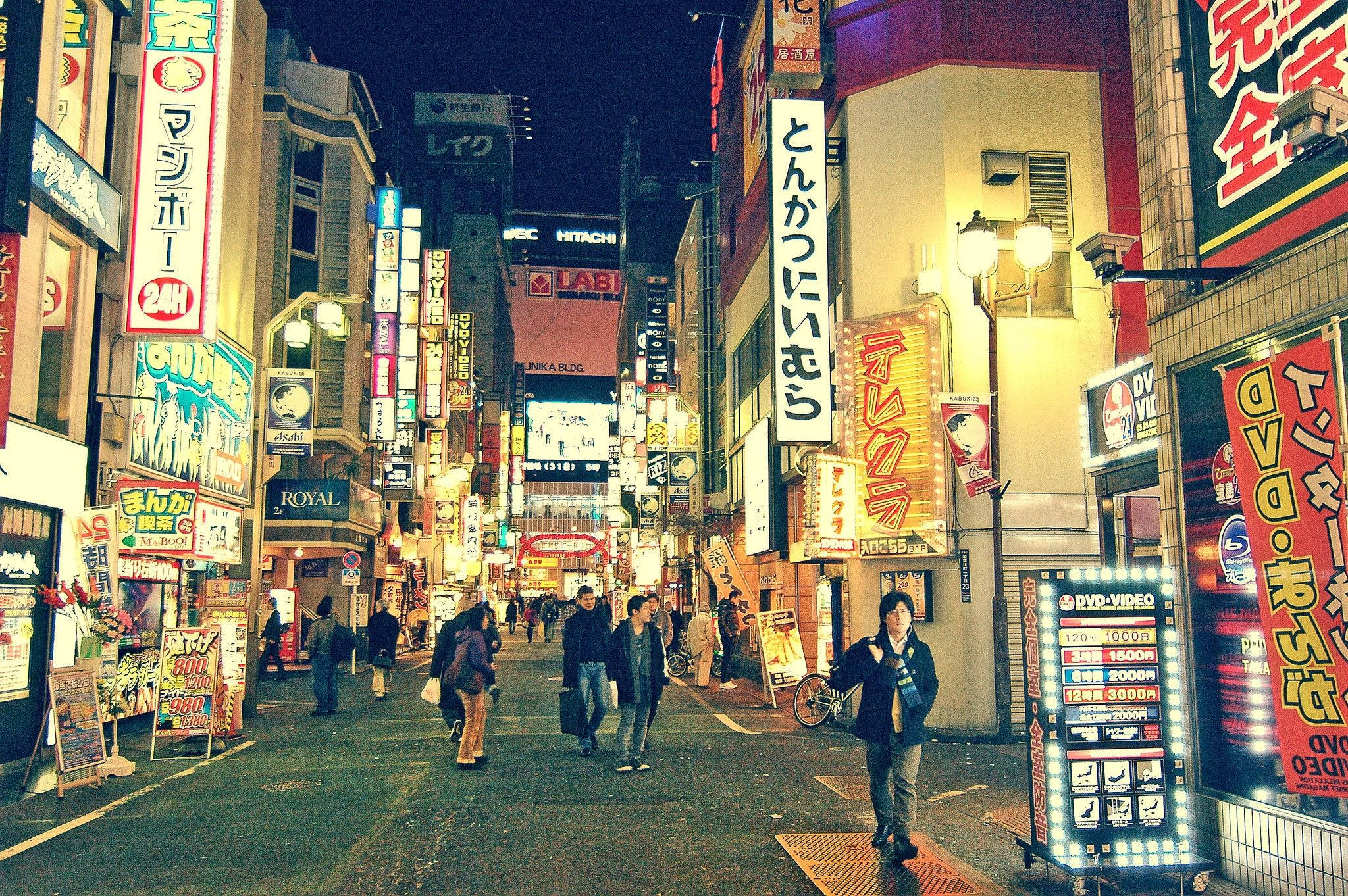 東京 桌布 壁紙 — 夜晚東京一條繁忙的霓虹燈街道，行人走過發光的招牌和明亮的店舖門面。