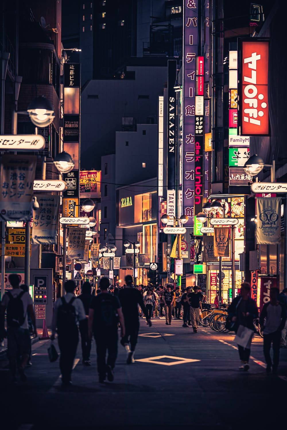 城市 桌布 壁紙 — 夜晚東京一條繁忙的霓虹燈街道，行人在發光的招牌和商店櫥窗下行走。