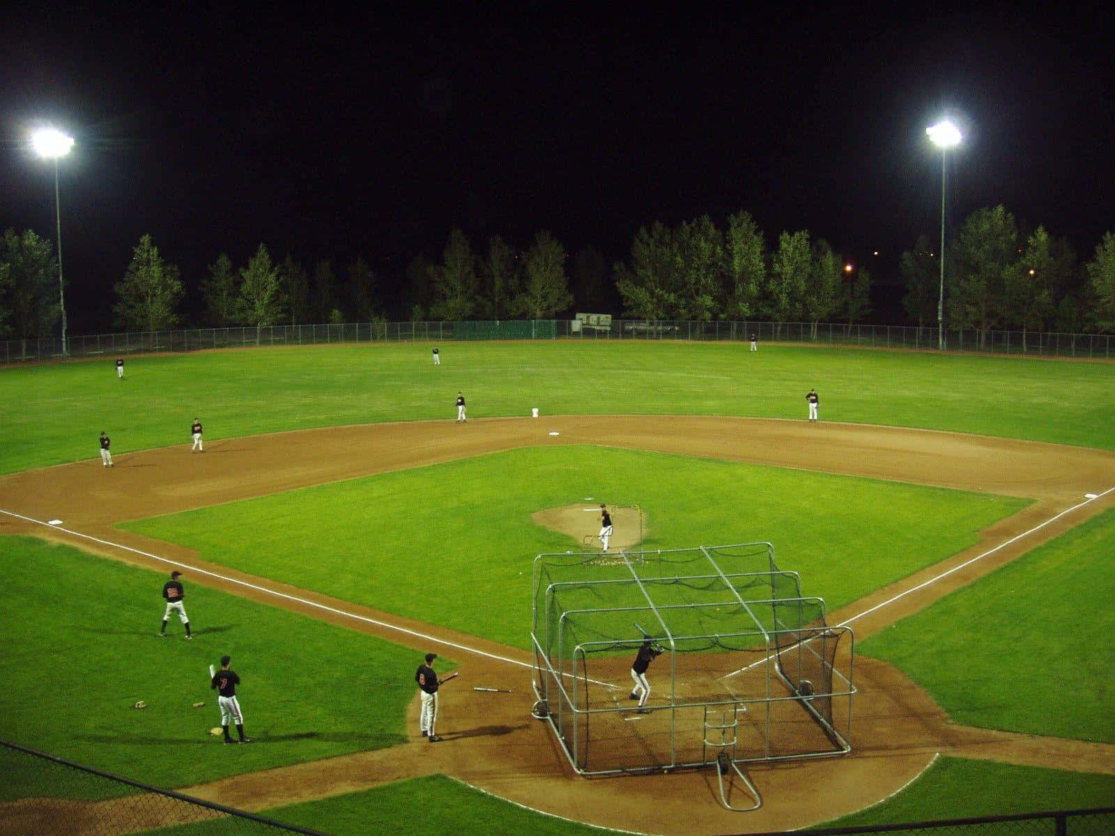 Baseball Field 桌布 壁紙 — 夜晚棒球比賽，在明亮的體育場燈光下，球員在光線充足的場地上，周圍環繞著樹木。