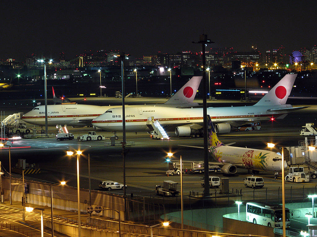 飛機 桌布 壁紙 — 夜晚機場停機坪，兩架日本航空波音747在明亮燈光下停泊在閘口，背景是城市天際線。
