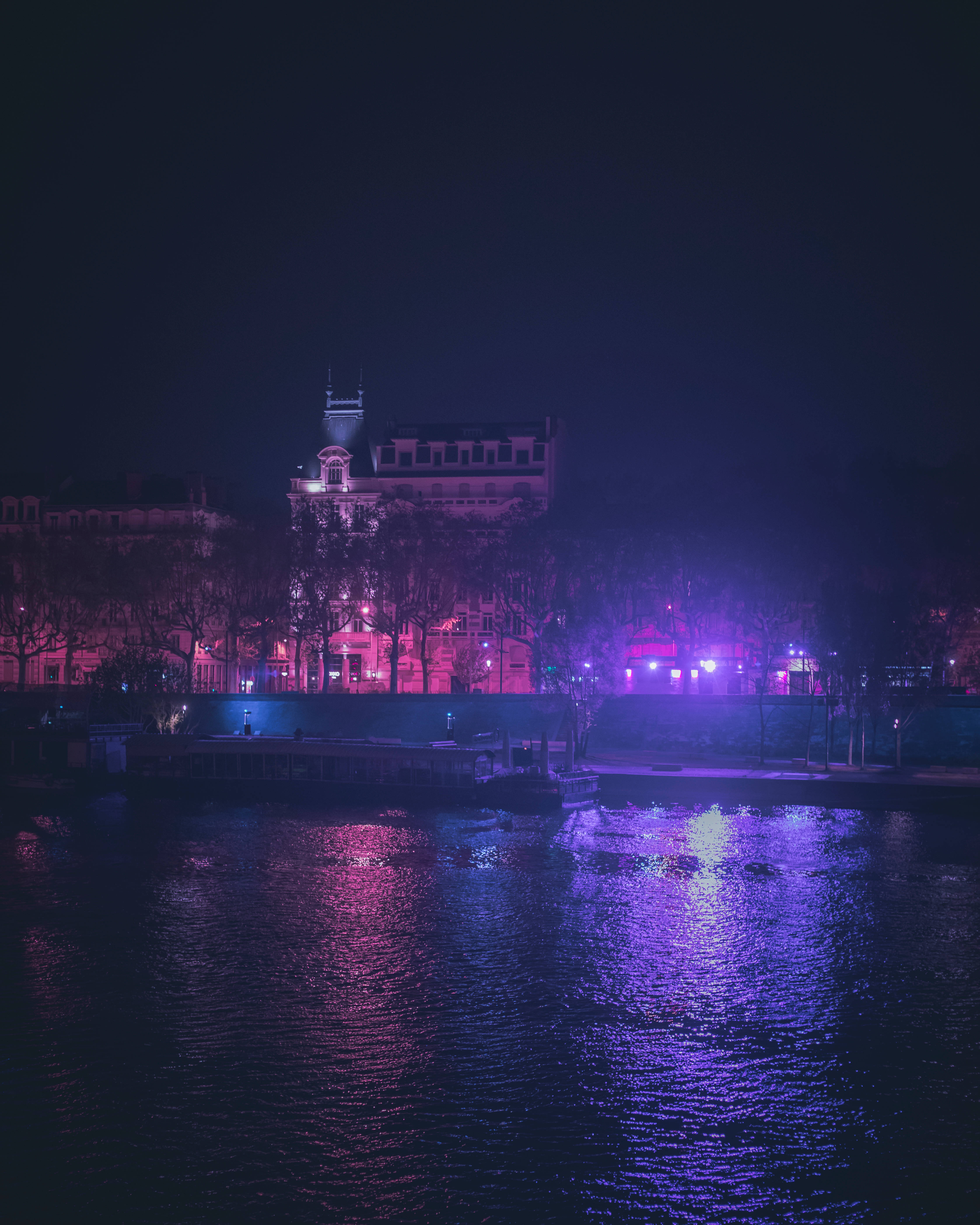 霓虹城市動態圖 桌布 壁紙 — 夜晚河邊景色，歷史建築物閃爍著粉紅和紫色燈光，倒映在漣漪的水面上。