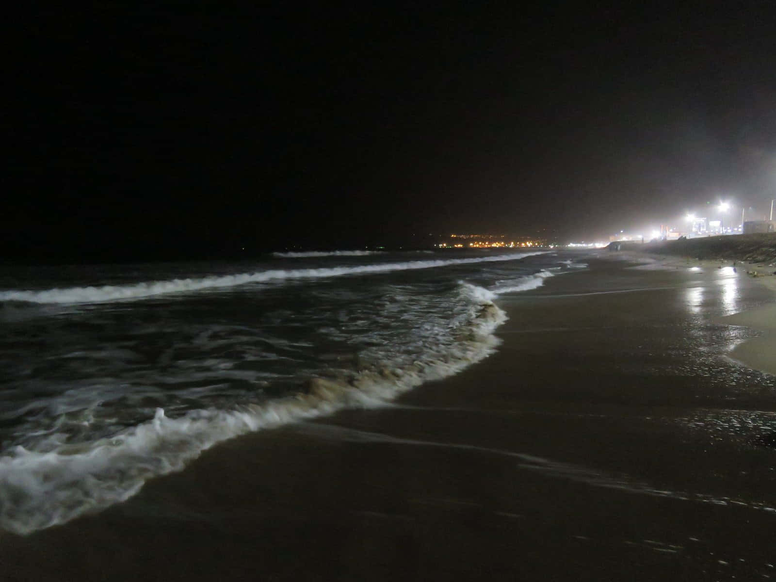 海灘 桌布 壁紙 — 夜晚海灘場景，海浪拍打岸邊，遠處城市燈光映照在濕沙上。