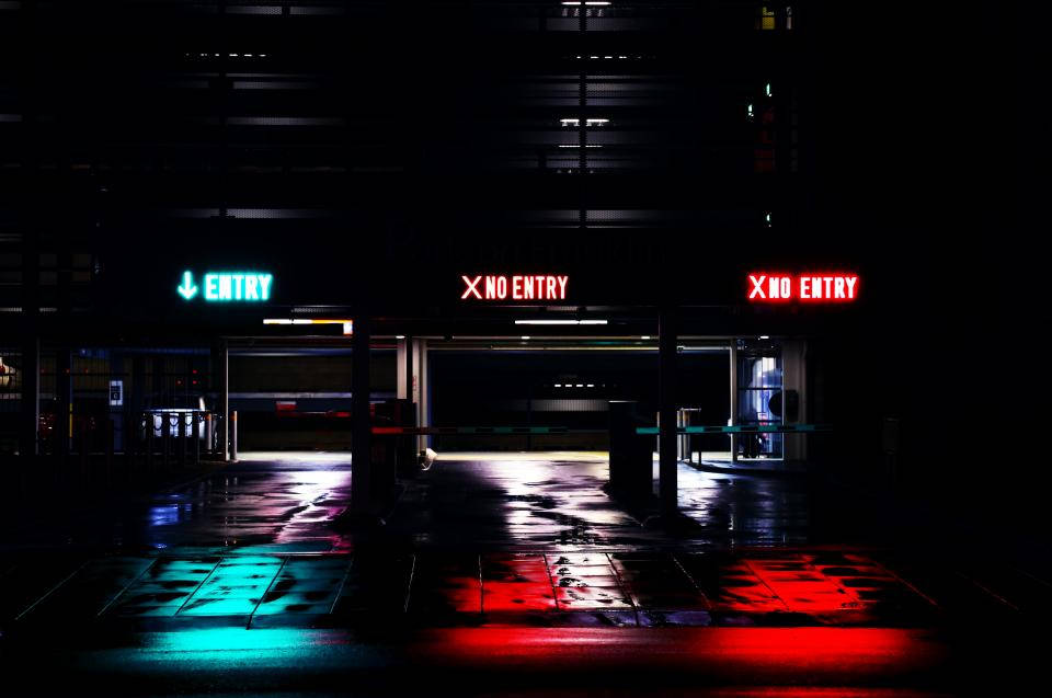 dark neon iPhone 桌布 壁紙 — 夜晚潮濕的停車場，發光的霓虹燈牌，一個綠色入口和兩個紅色禁止進入，在地板上投射出色彩繽紛的倒影。