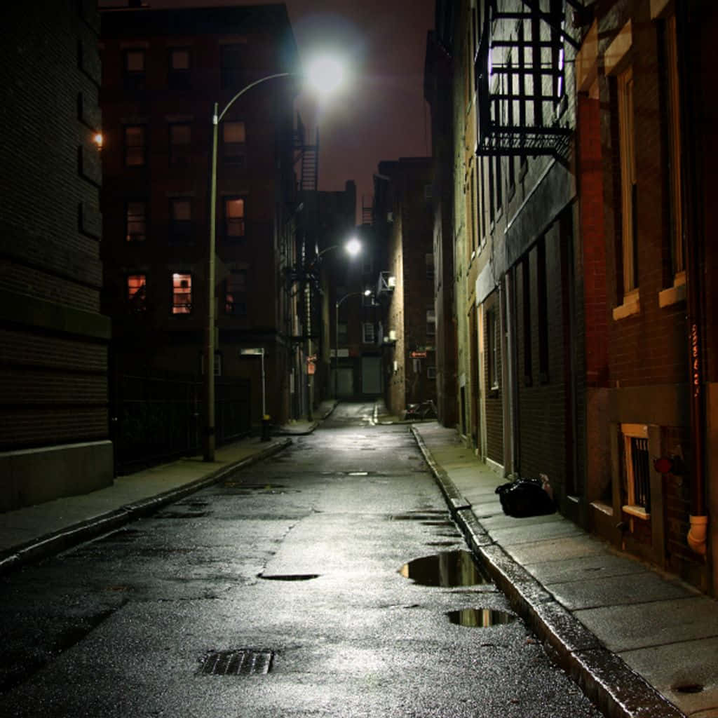 Dark Alleyway 桌布 壁紙 — 夜晚潮濕的城市小巷，街燈照亮並反射在磚樓之間的水坑中。