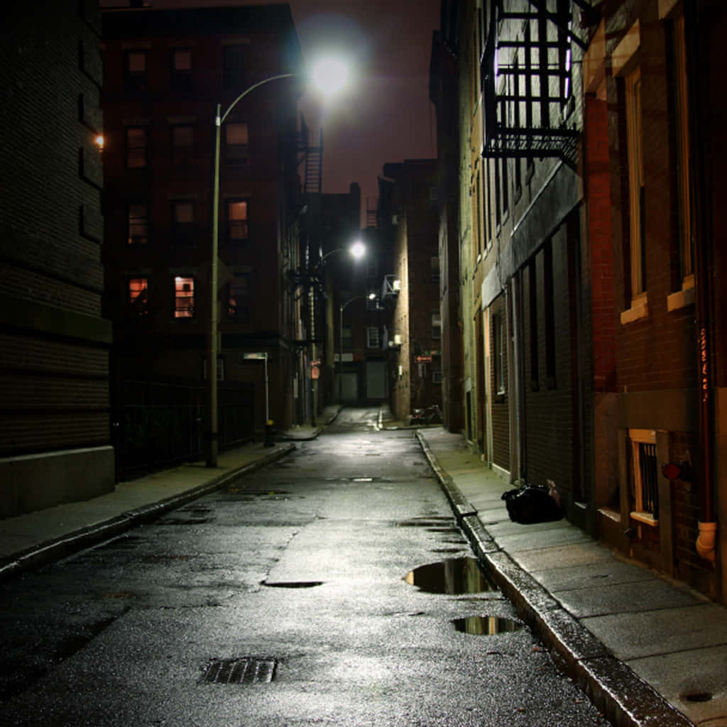 Alleyway 桌布 壁紙 — 夜晚潮濕的城市小巷，街燈照亮並反射在磚樓之間的水坑中。