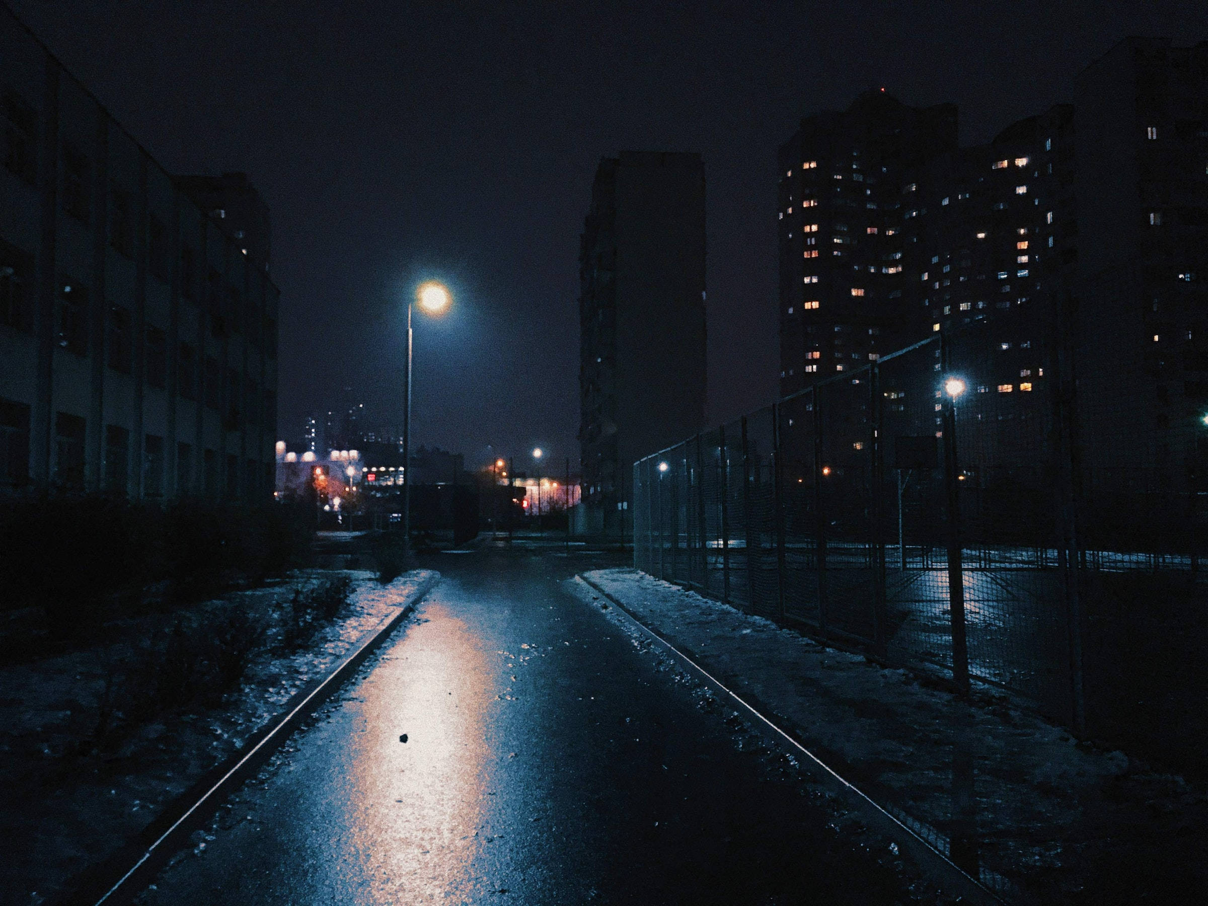 夜空 桌布 壁紙 — 夜晚濕漉漉的城市小巷，在黑暗的天空下反射著街燈，地面覆蓋著雪，背景是高樓大廈。