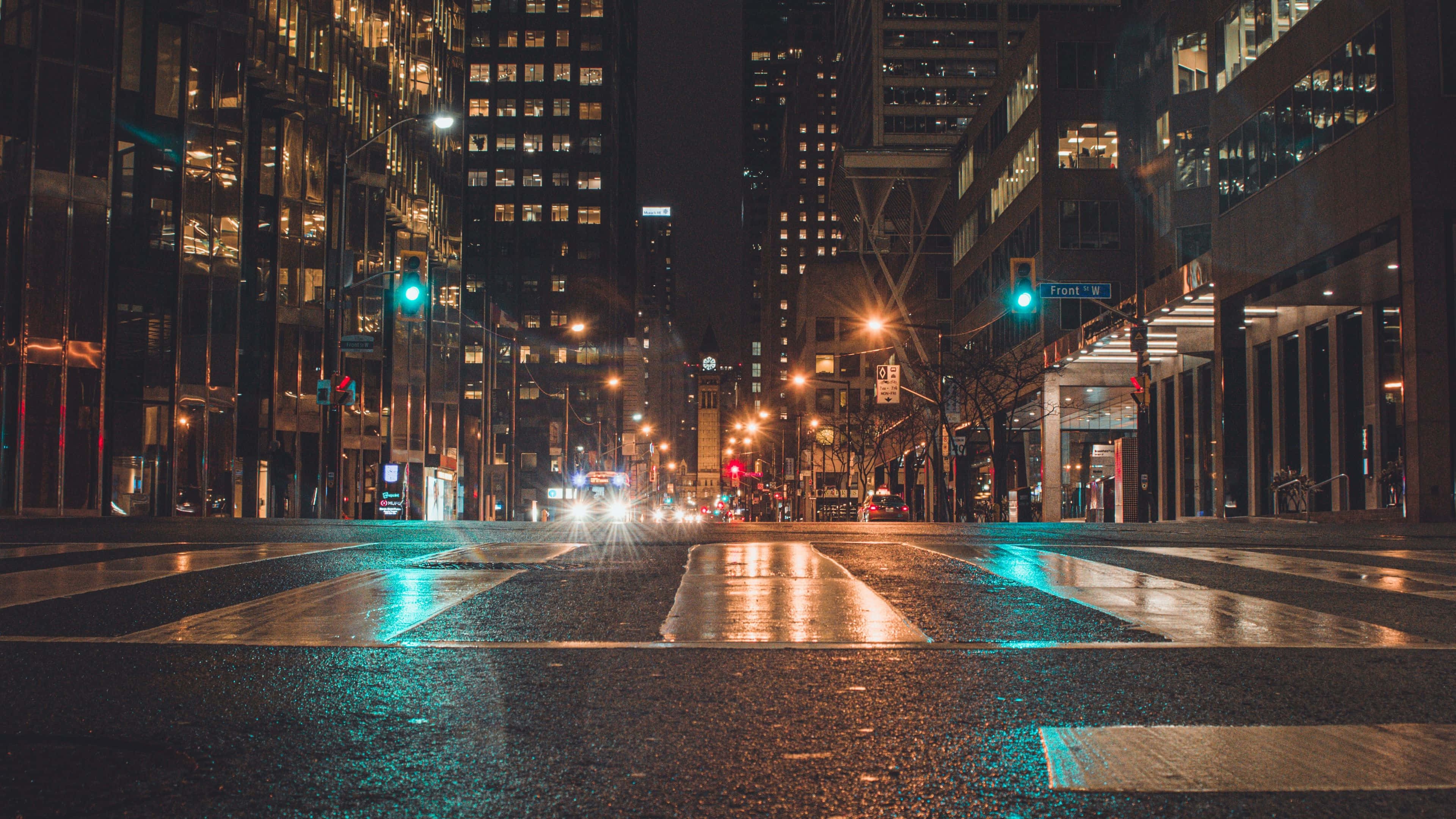 夜景城市 桌布 壁紙 — 夜晚濕漉漉的城市街道，在高聳的玻璃建築之間反射著閃爍的路燈和交通信號燈。