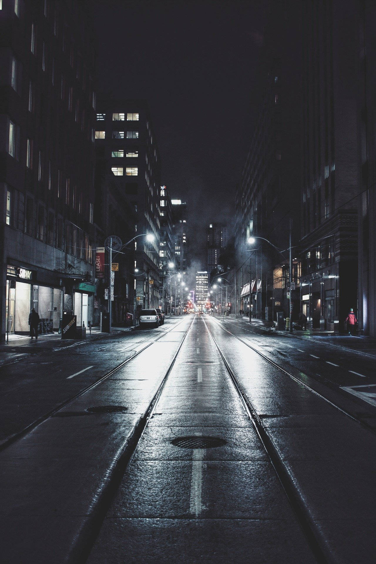 深色 iPhone 桌布 壁紙 — 夜晚濕漉漉的城市街道反射出路燈和建築物窗戶，營造出陰鬱的城市峽谷效果。