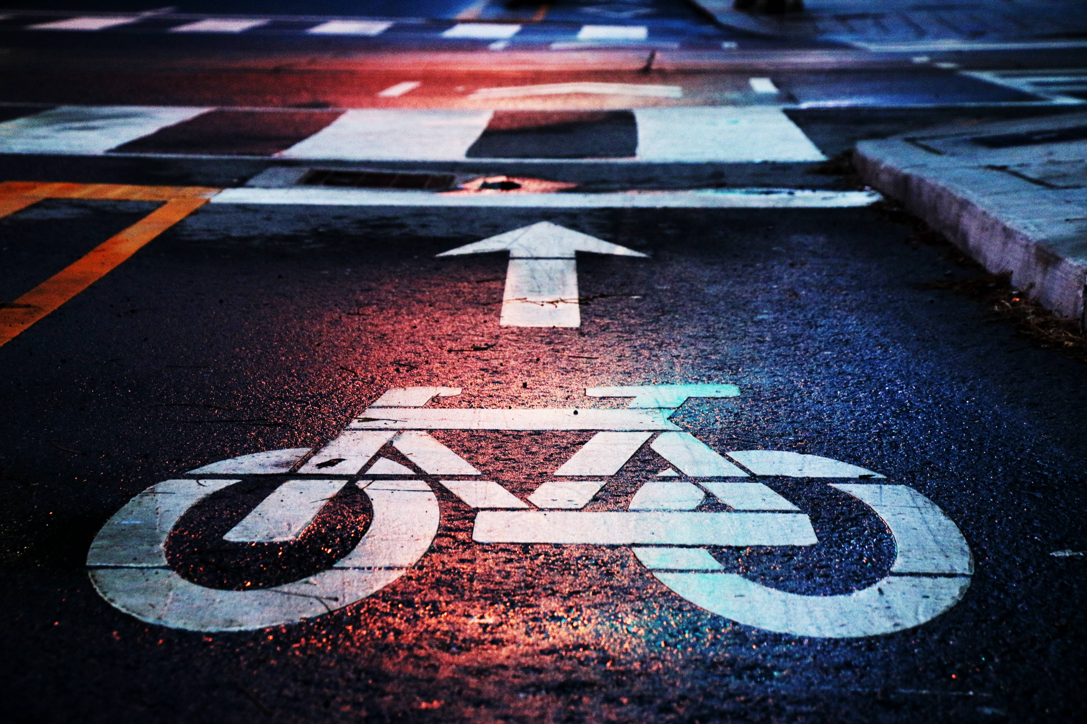 Bicycle 桌布 壁紙 — 夜晚濕瀝青路面的特寫，帶有褪色的白色自行車道標誌和箭頭，被鮮豔的紅色和藍色街燈照亮。