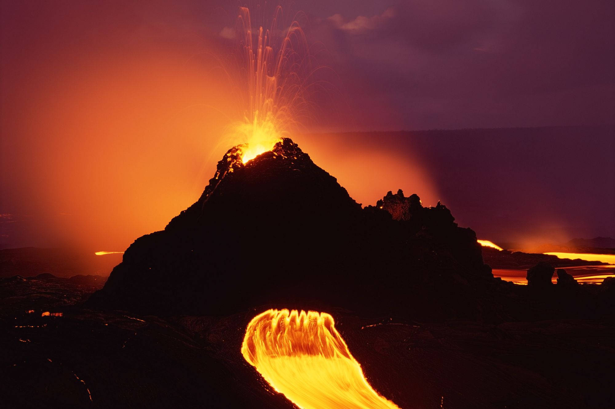Volcano 桌布 壁紙 — 夜晚火山爆發，發光的熔岩噴泉和流動的熔岩河流映襯著黑暗的天空。