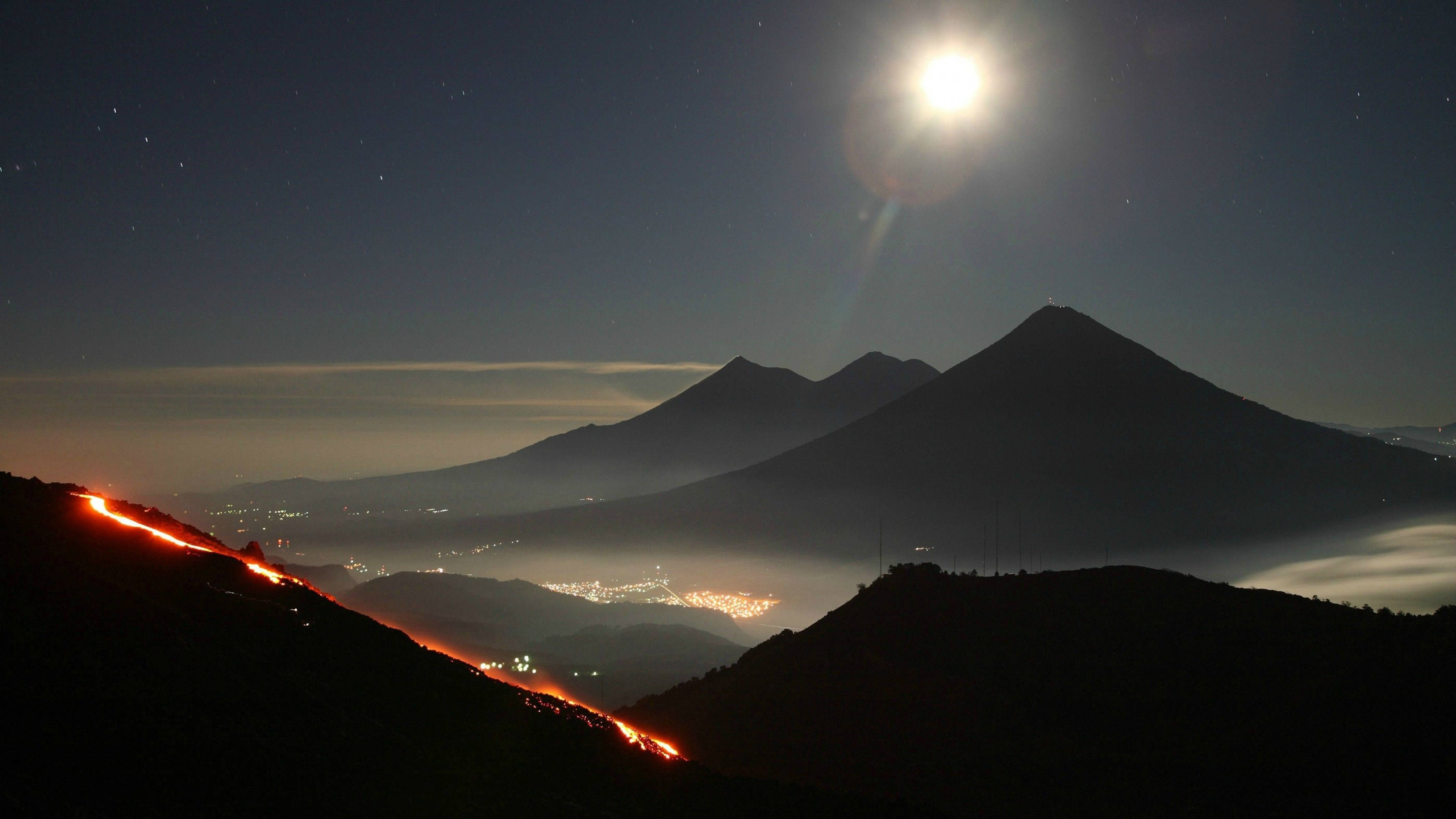 Guatemala 桌布 壁紙 — 夜晚火山爆發，發光的熔岩流在明亮的月亮和星空下照亮黑暗的山脈。
