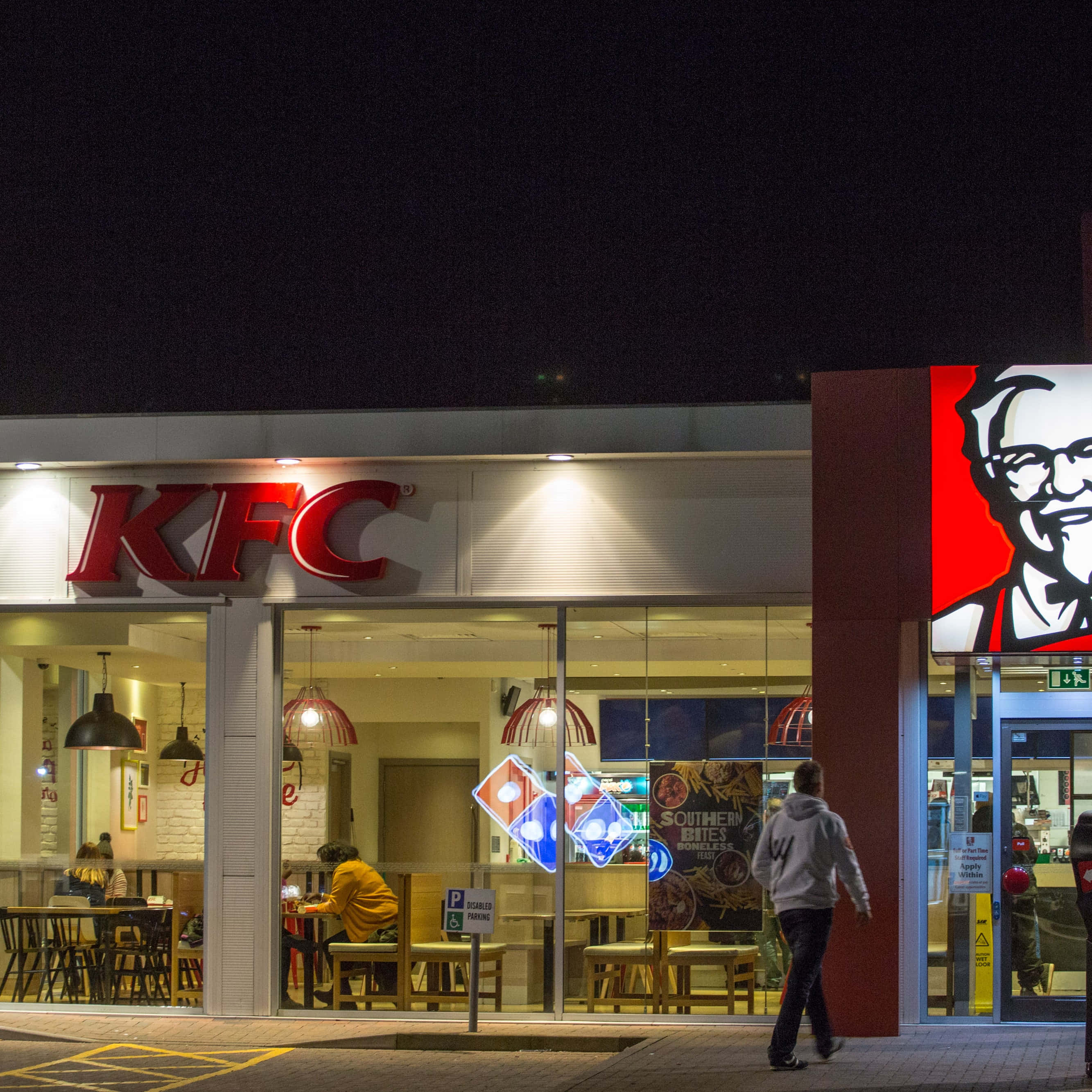 Kfc 桌布 壁紙 — 夜晚燈光明亮的KFC餐廳，發光的招牌和一個人走過入口。