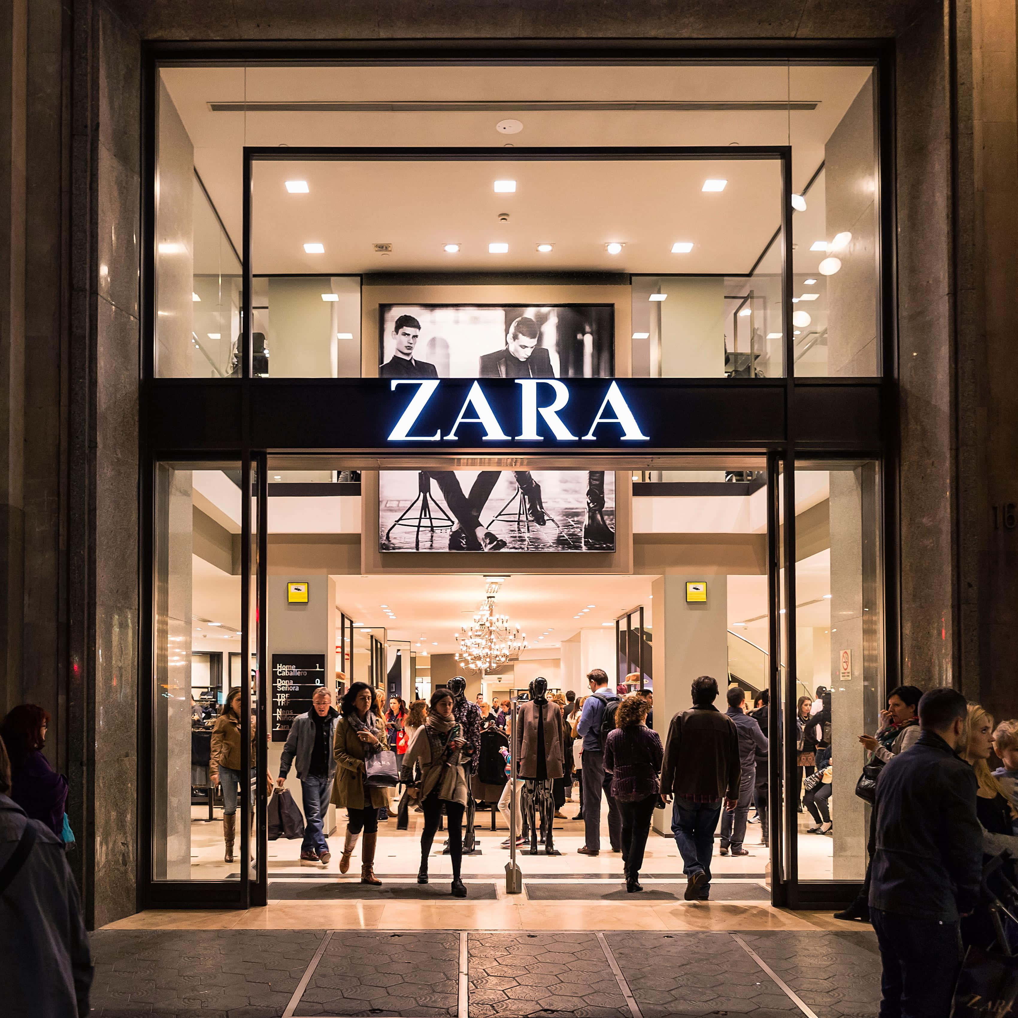 Zara 桌布 壁紙 — 夜晚燈光明亮的Zara店舖入口，人們通過玻璃門進出。