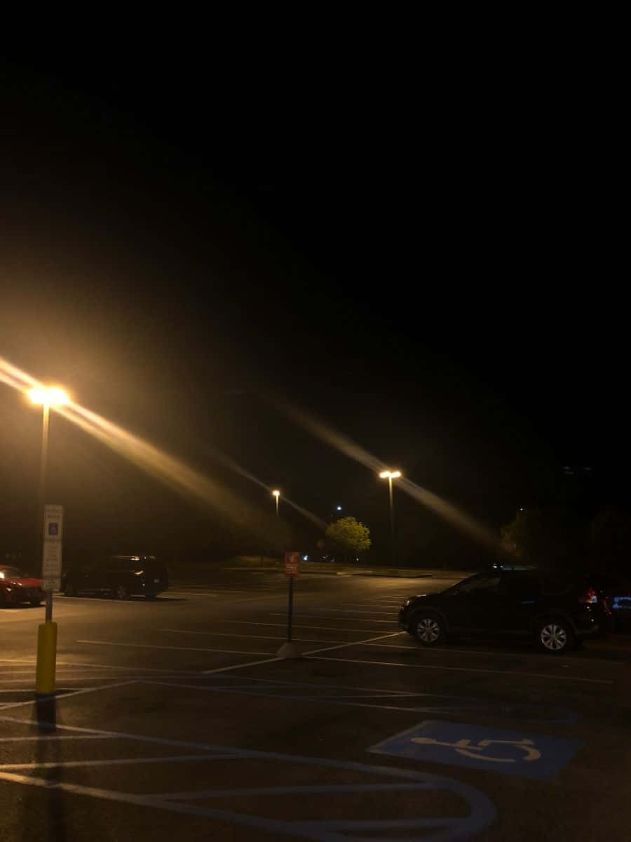 Night Aesthetic 桌布 壁紙 — 夜晚燈光昏暗的停車場，街燈在停泊的汽車和標記的車位上投下長長的光束。