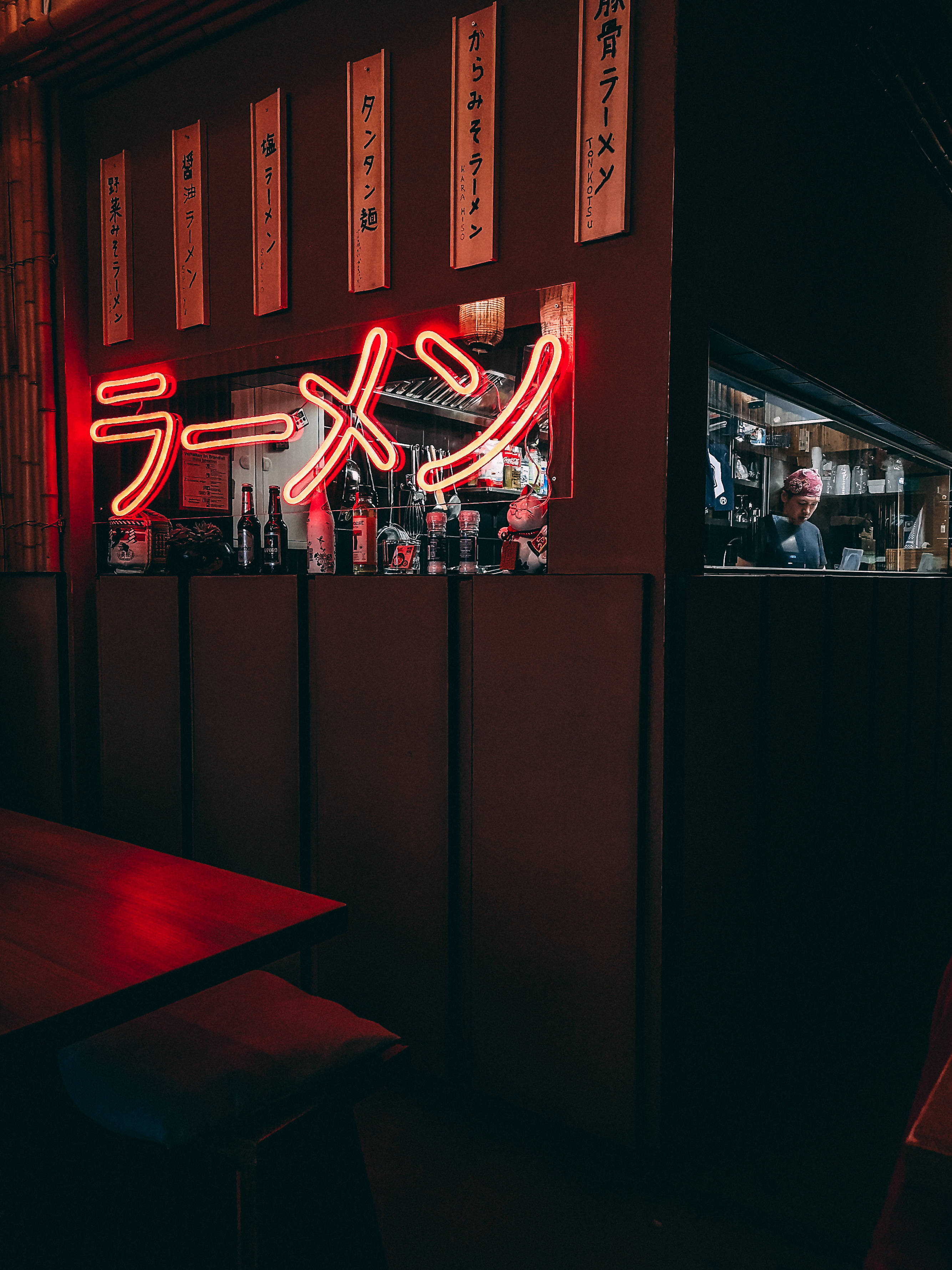 Neon Phone 桌布 壁紙 — 夜晚燈光昏暗的拉麵吧，發光的紅色霓虹燈招牌在木桌上投下溫暖的光線，櫃檯後方可見廚師。