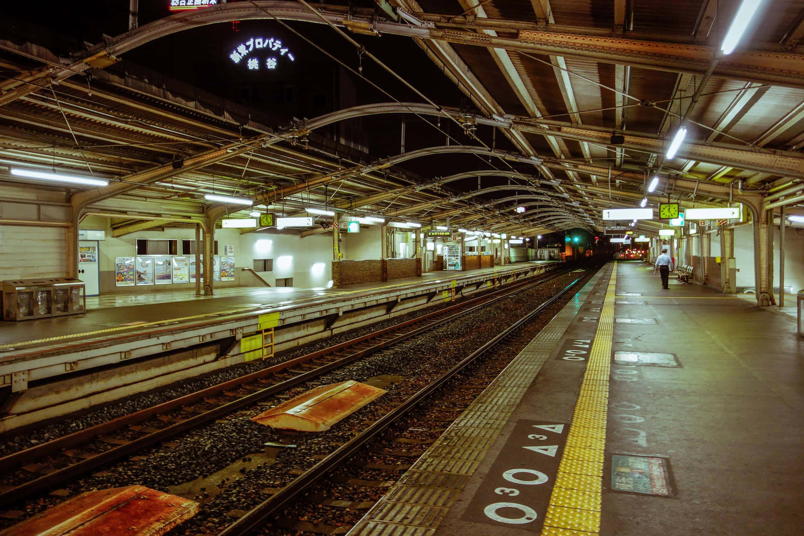 Train Station 桌布 壁紙 — 夜晚燈光昏暗的火車站月台，頭頂燈光在軌道上投下溫暖的光芒，一名孤獨的乘客正在走遠。