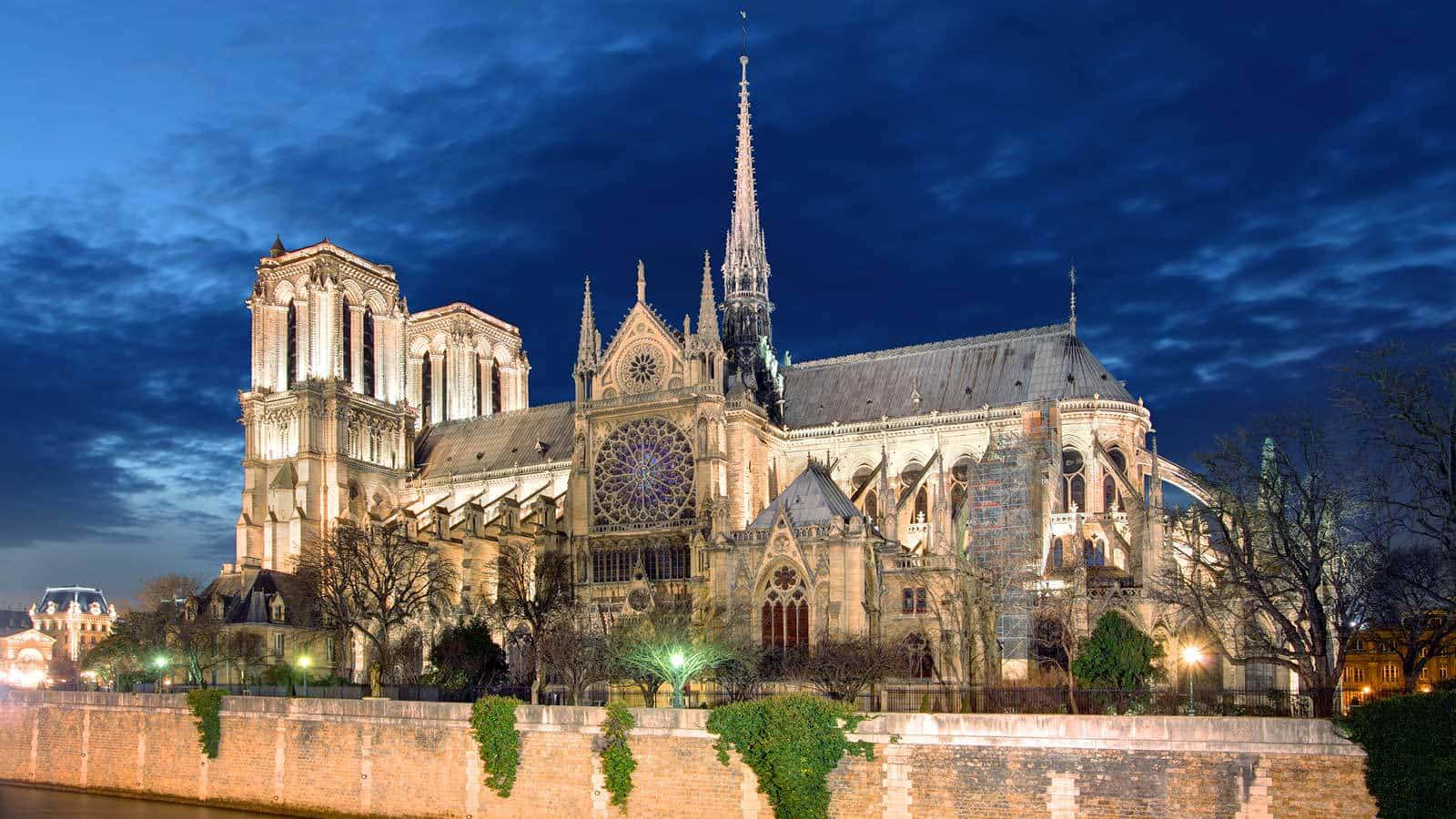 Notre Dame 桌布 壁紙 — 夜晚燈光照耀下的巴黎聖母院，背景是深藍色的天空和戲劇性的雲彩。