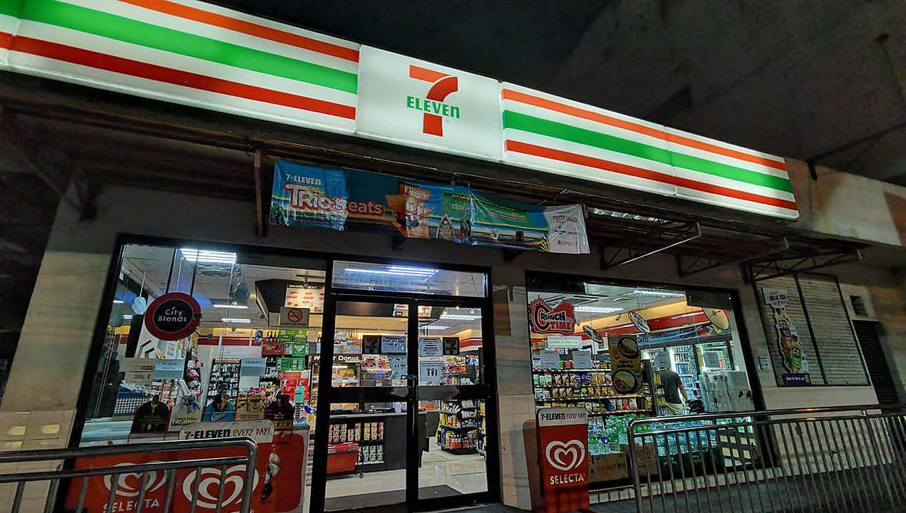 7-ELEVEN 桌布 壁紙 — 夜晚燈火通明的7-Eleven店鋪，色彩繽紛的招牌和玻璃門展示內部貨架上的商品。