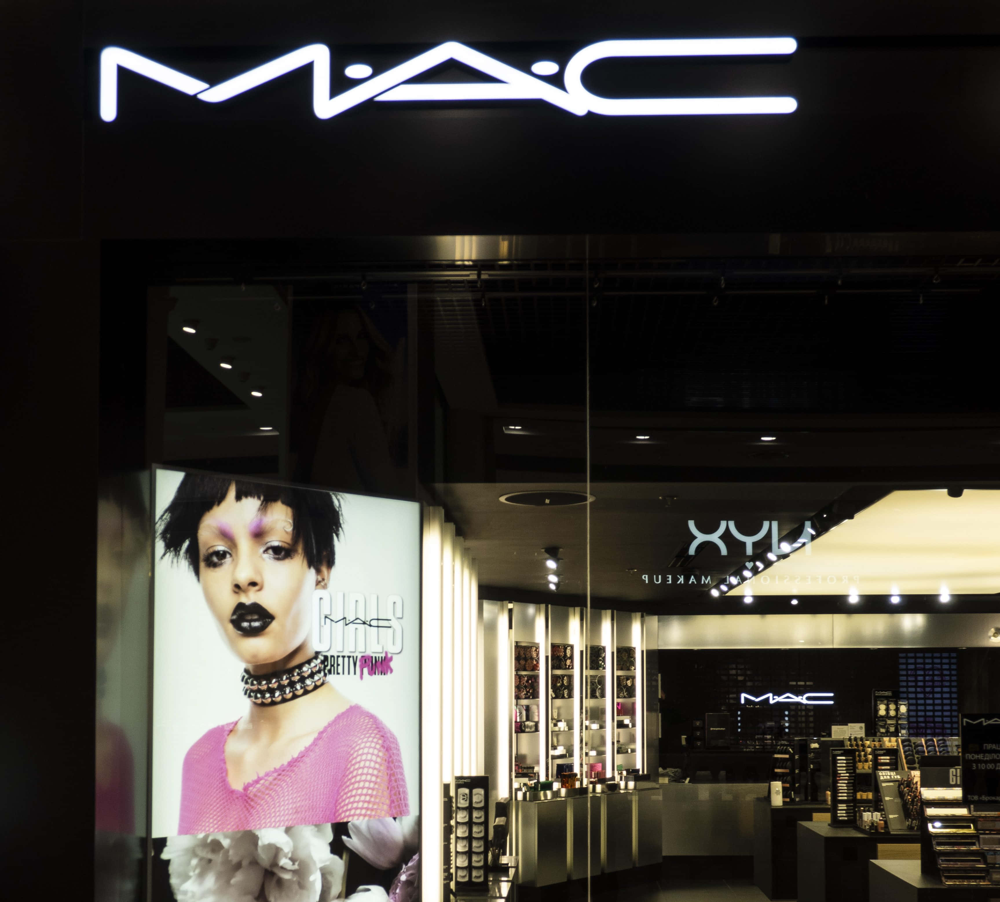 Mac Cosmetics 桌布 壁紙 — 夜晚燈火通明的MAC化妝品店，有一張大海報，模特兒化著大膽妝容，身穿粉紅色服裝。