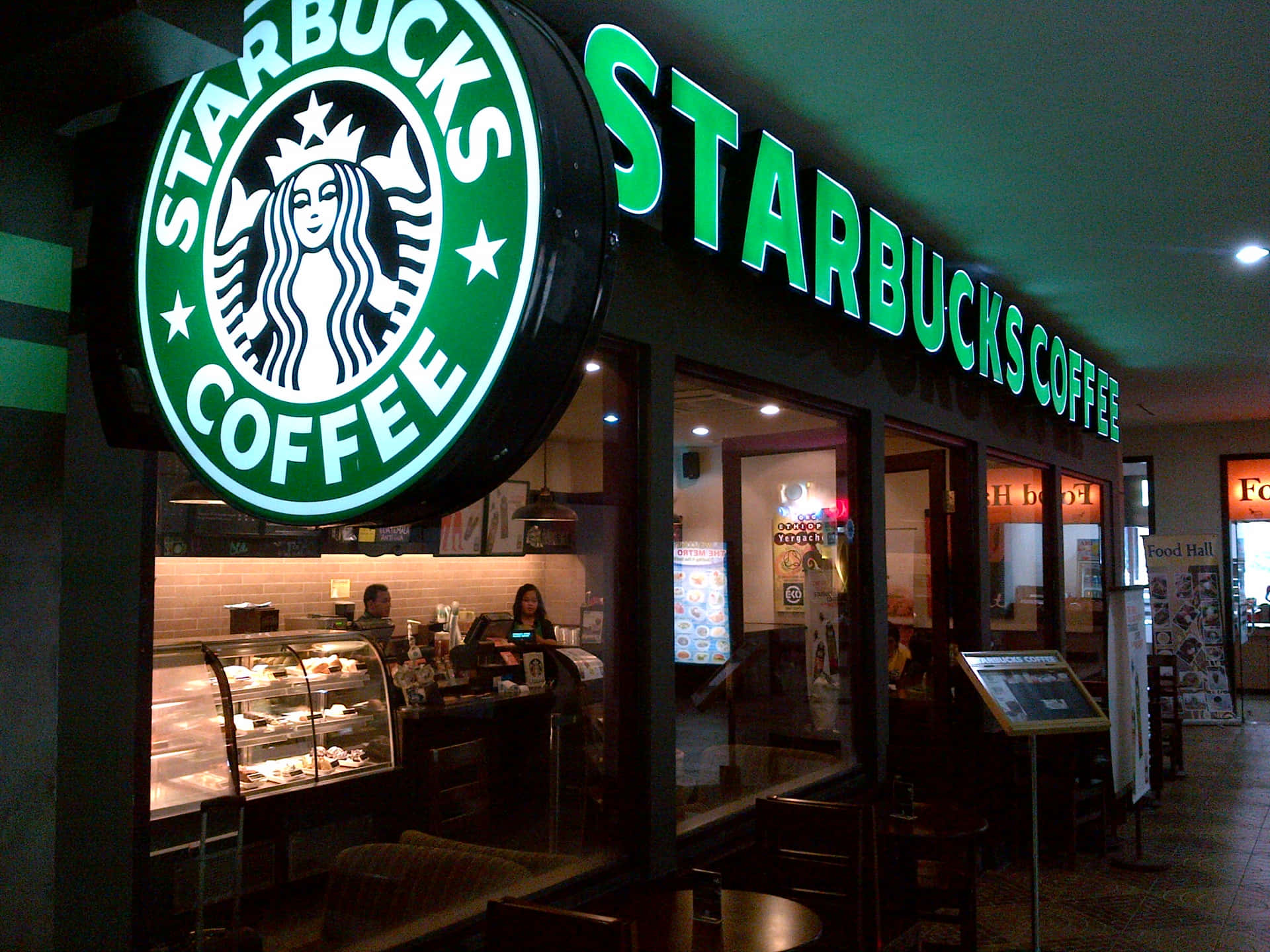 星巴克 桌布 壁紙 — 夜晚燈火通明的Starbucks咖啡店，帶有發光的綠色招牌，內部座位和展示櫃清晰可見。