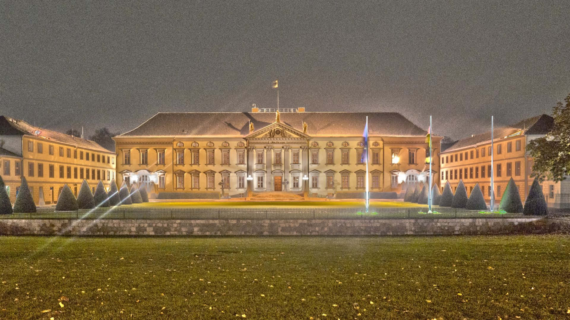 Berlin 桌布 壁紙 — 夜晚燈火通明的新古典主義宮殿，兩翼對稱，旗幟飄揚，前景有修剪整齊的圓錐形樹籬。