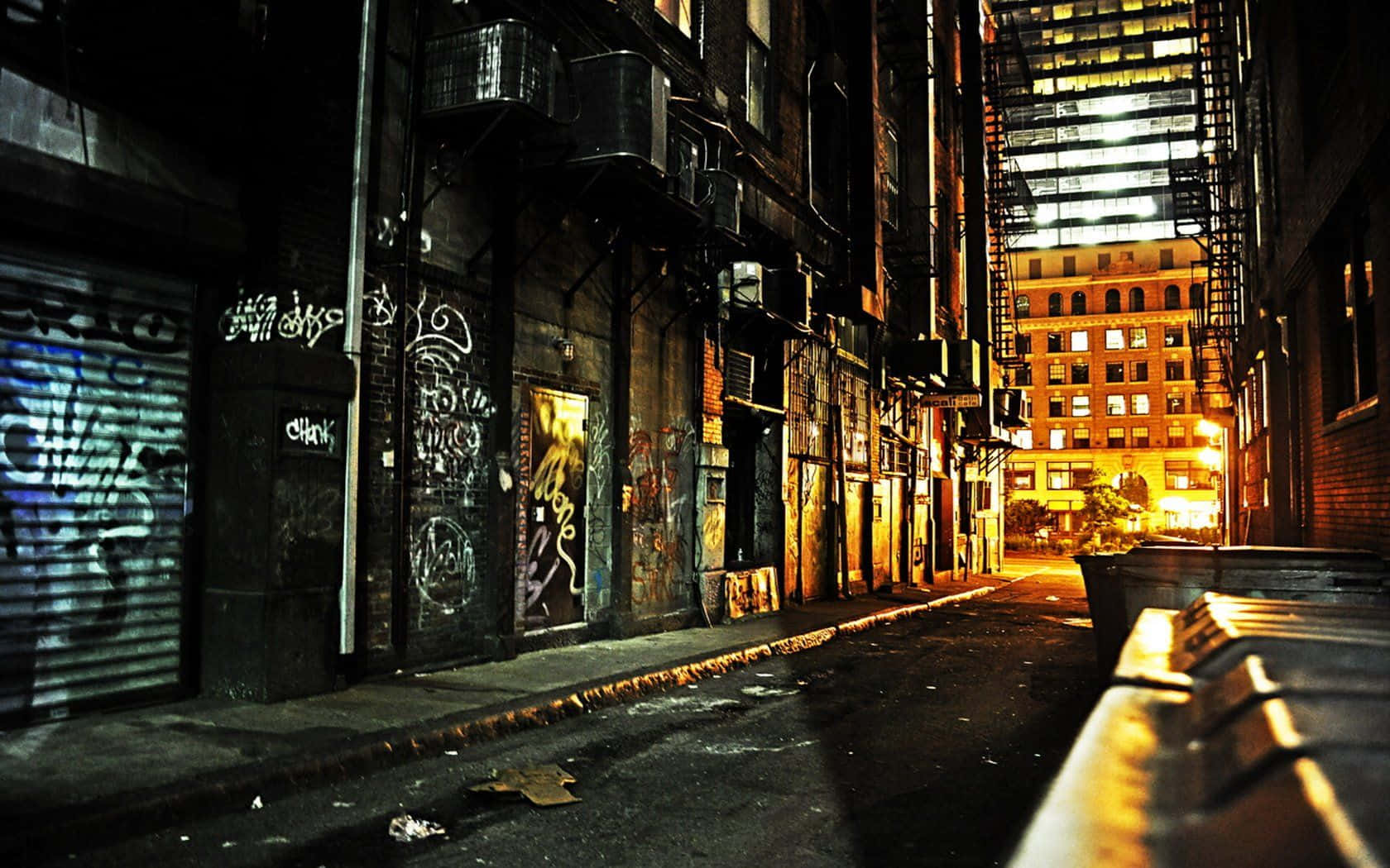 Dark Alleyway 桌布 壁紙 — 夜晚狹窄的城市小巷，溫暖的路燈照亮，牆上佈滿塗鴉，遠處有明亮的建築物。