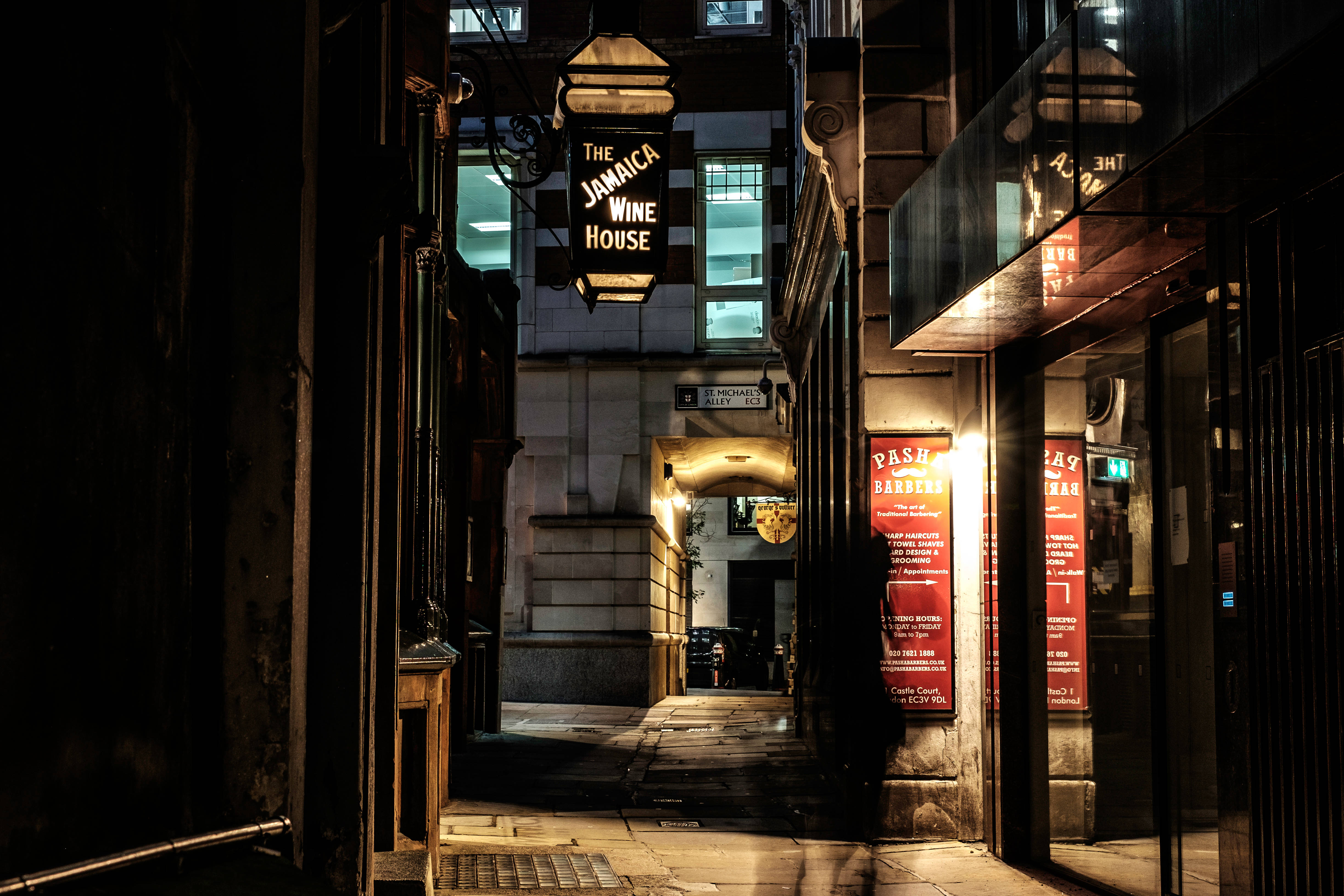 Street 桌布 壁紙 — 夜晚狹窄的城市小巷，被'The Jamaica Wine House'溫暖發光的招牌和紅色海報照亮，在石牆上投下長長的陰影。