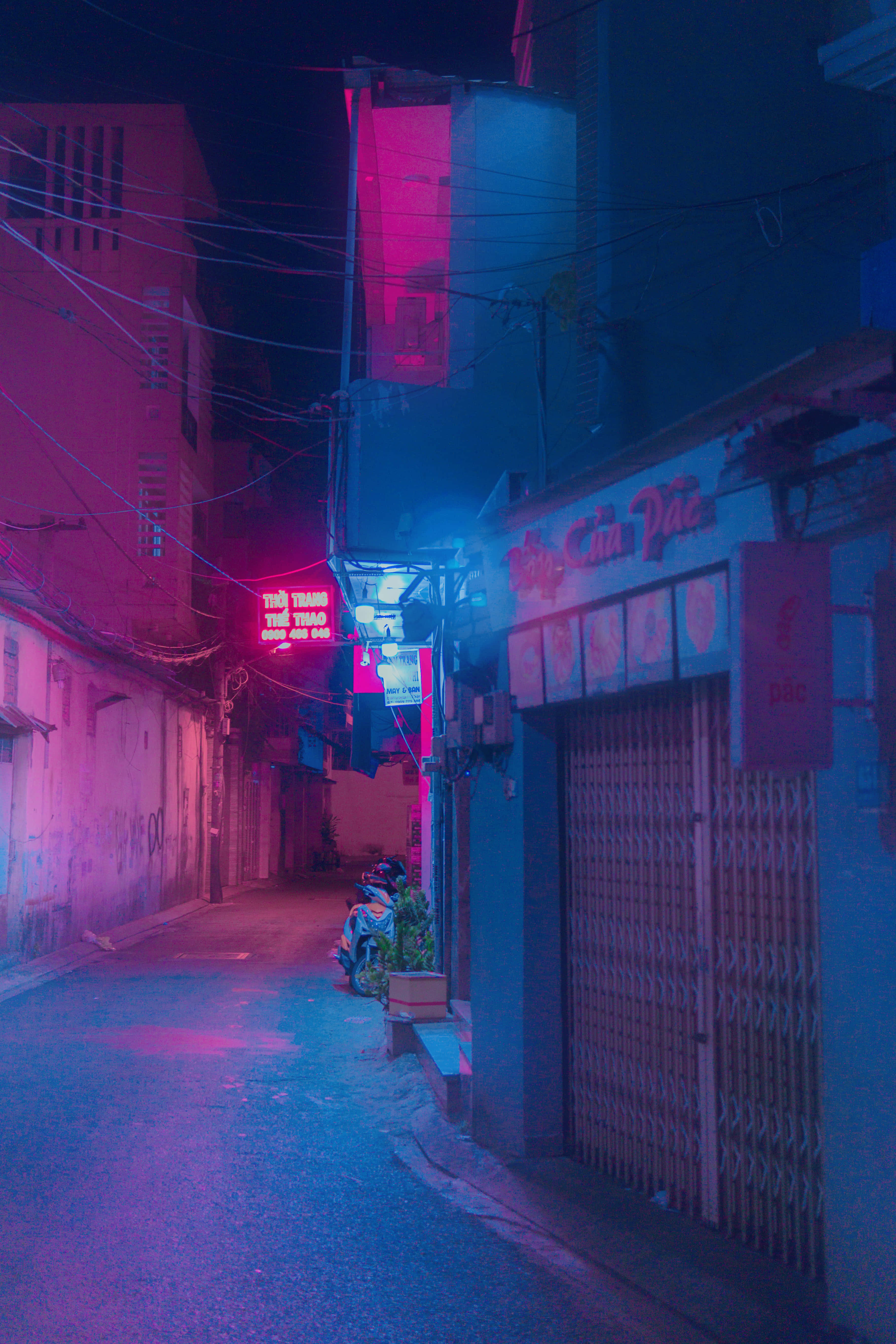 Neon Aesthetic Phone 桌布 壁紙 — 夜晚狹窄的城市小巷，被發光的招牌和關閉的商店百葉窗投射出的鮮豔粉紅和藍色霓虹燈光所沐浴。