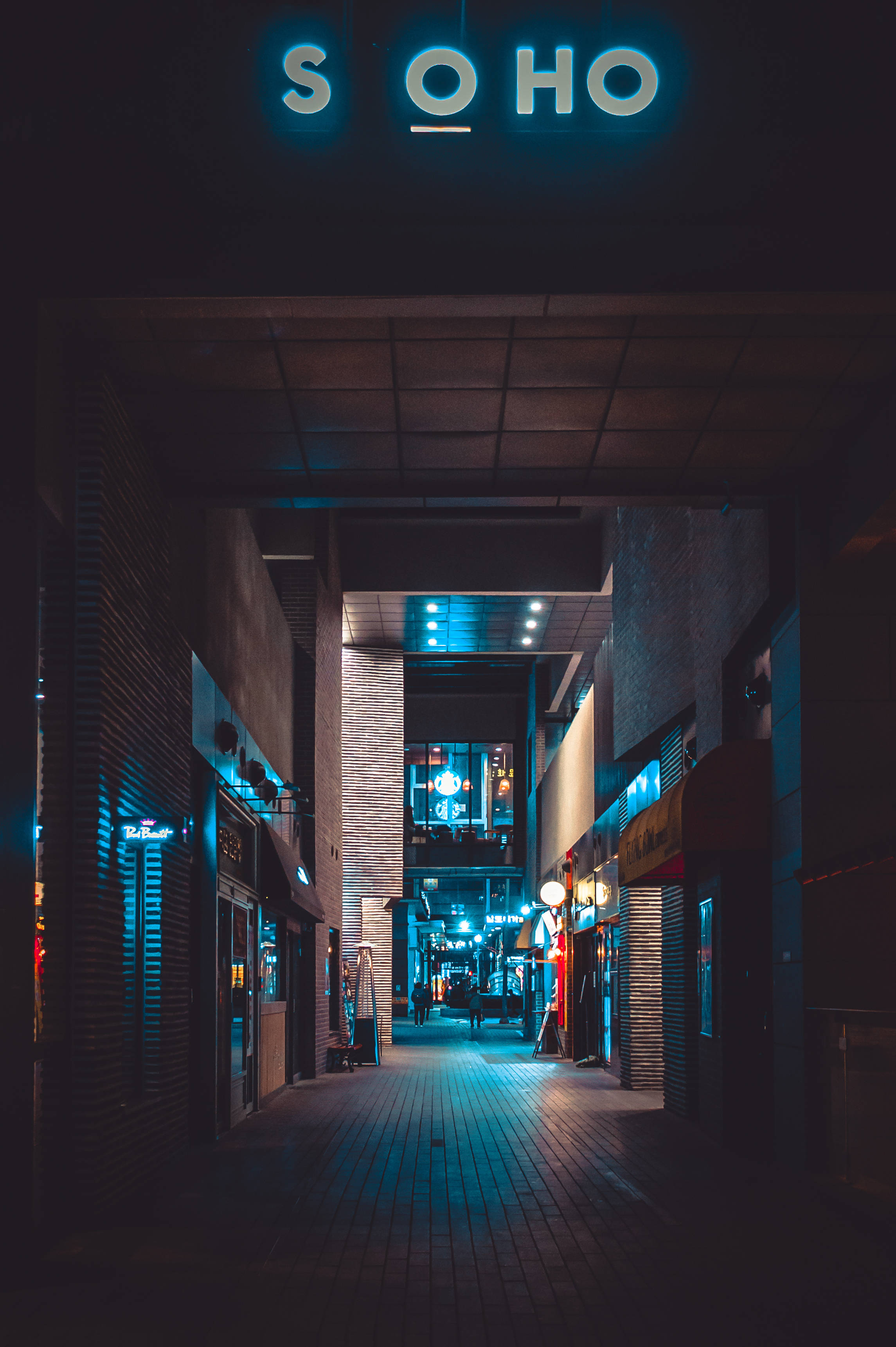 dark neon iPhone 桌布 壁紙 — 夜晚狹窄的城市小巷，被發光的藍色霓虹燈招牌和溫暖的路燈照亮，投下長長的影子。