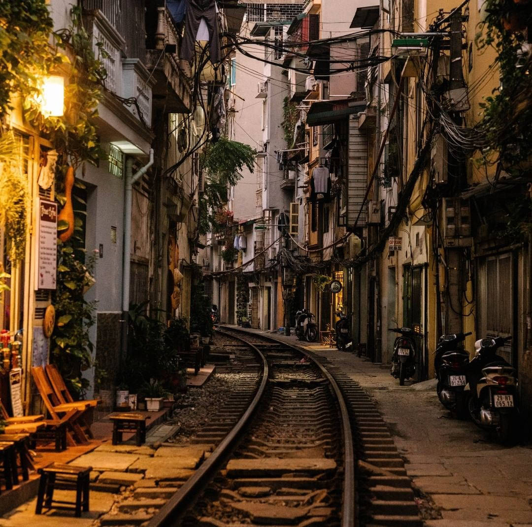 河內 桌布 壁紙 — 夜晚狹窄的城市小巷，鐵路軌道蜿蜒穿過，兩旁建築物掛著晾曬的衣物，暖色街燈下停著機車。