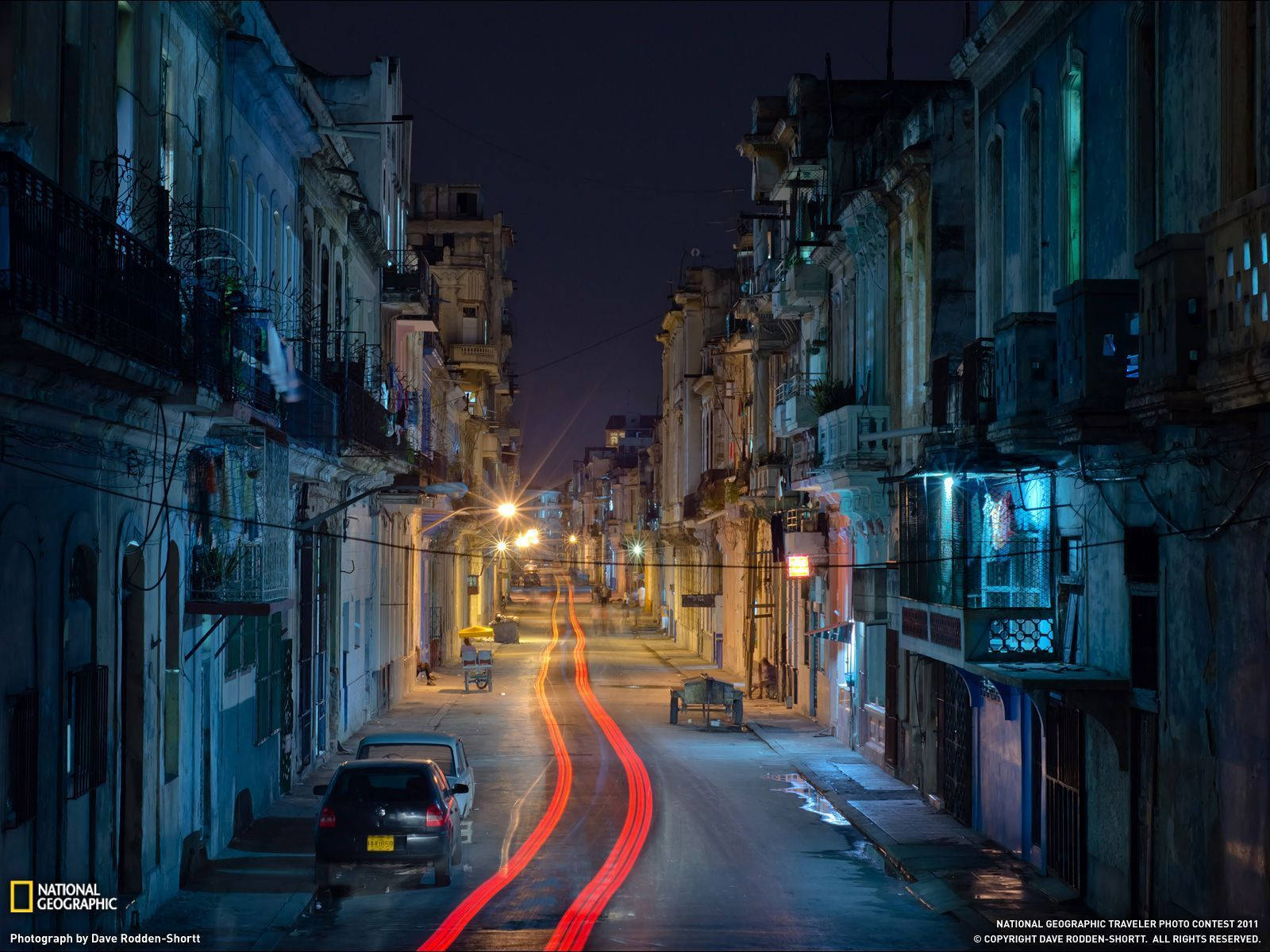 Cuba 桌布 壁紙 — 夜晚狹窄的城市街道，經過風化的建築物之間有汽車駛過時留下的紅色光跡。