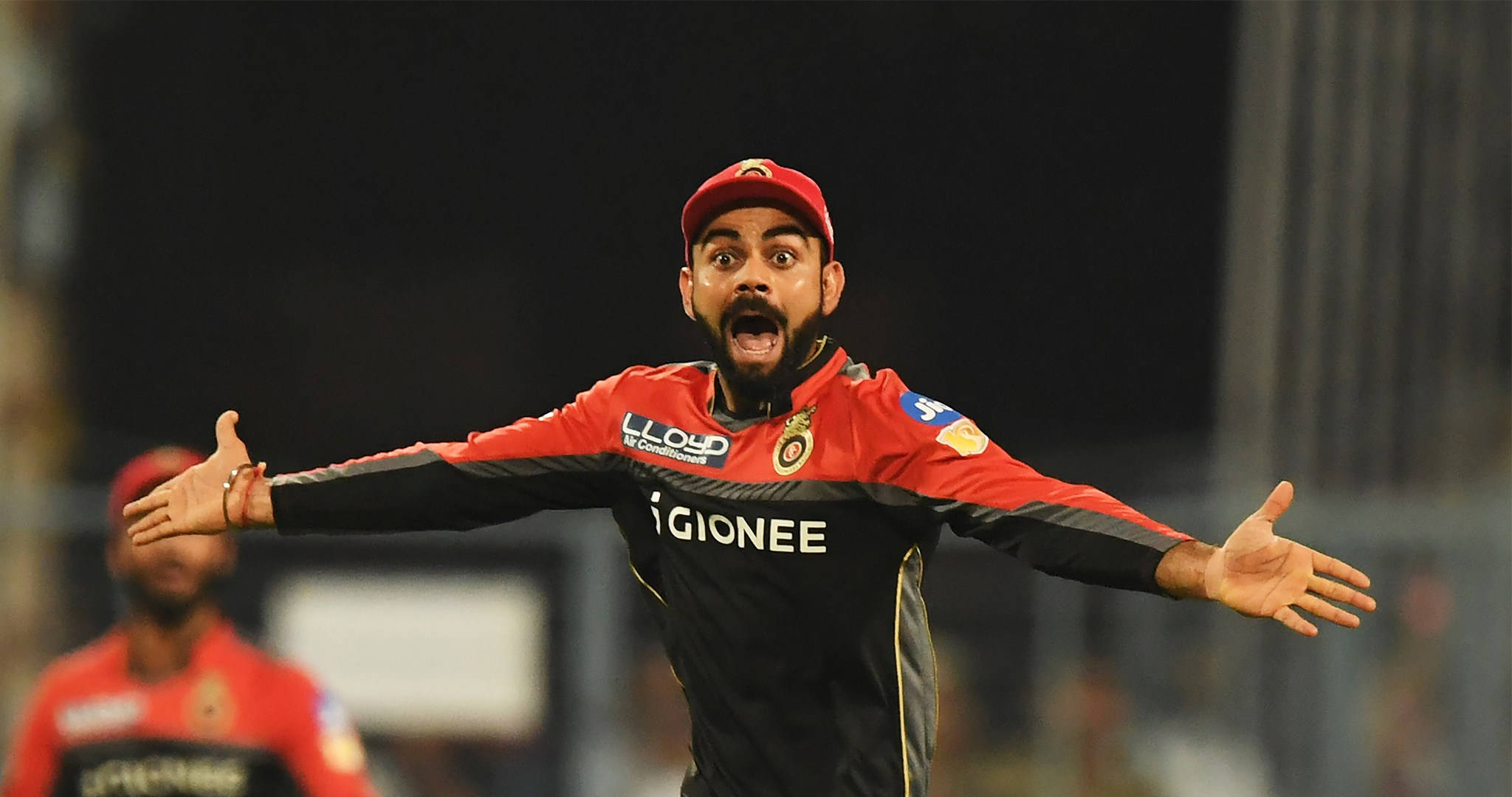 royal challengers bangalore 桌布 壁紙 — 夜晚球場燈光下，戴紅帽穿紅球衣的板球運動員張開雙臂慶祝。