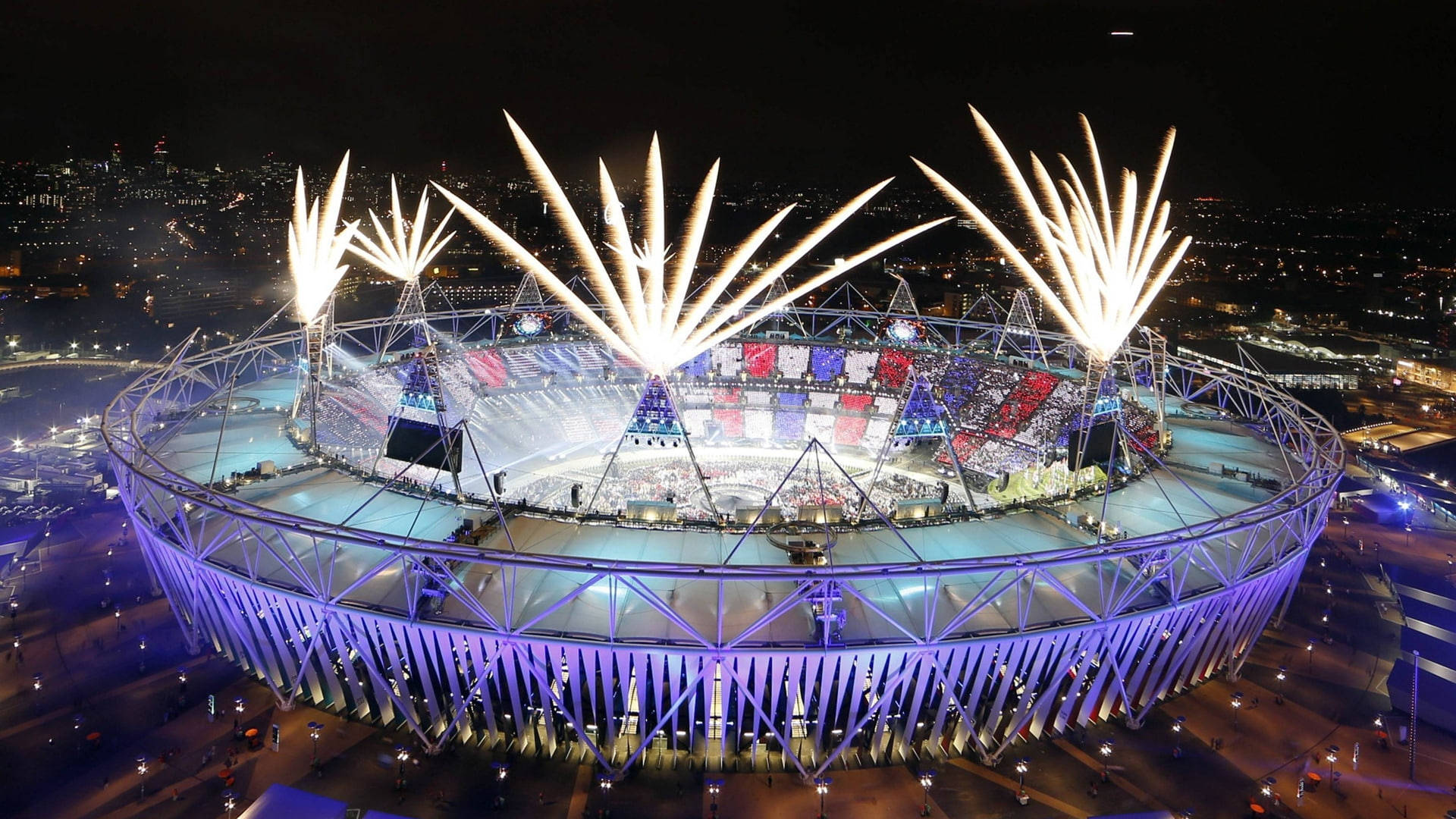 Olympics 桌布 壁紙 — 夜晚發光的奧林匹克體育場鳥瞰圖，煙火在人群上方爆炸。