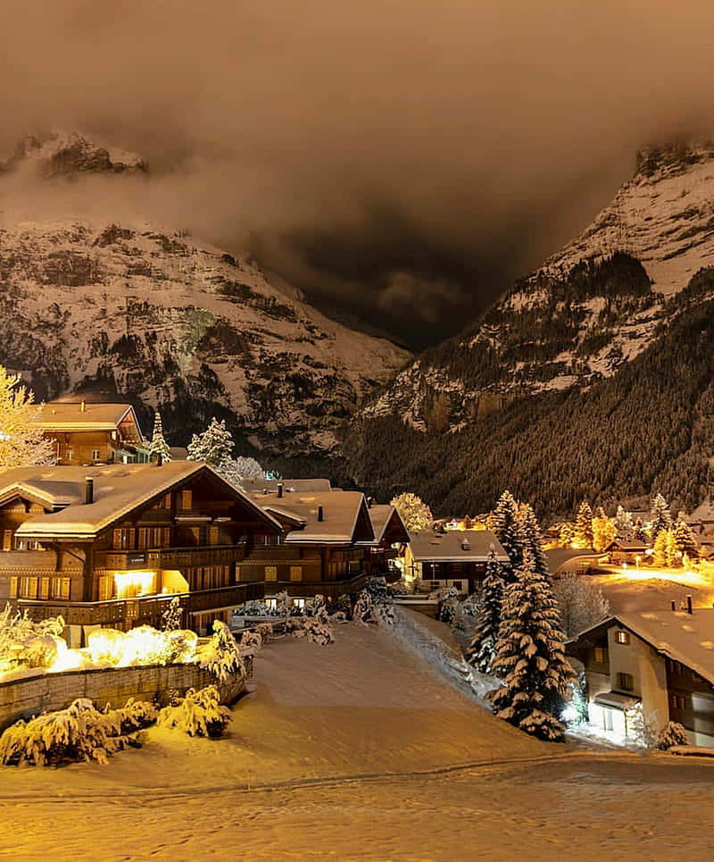 Christmas Winter Wonderland 桌布 壁紙 — 夜晚白雪覆蓋的山村，溫暖發光的窗戶和松樹在烏雲下。