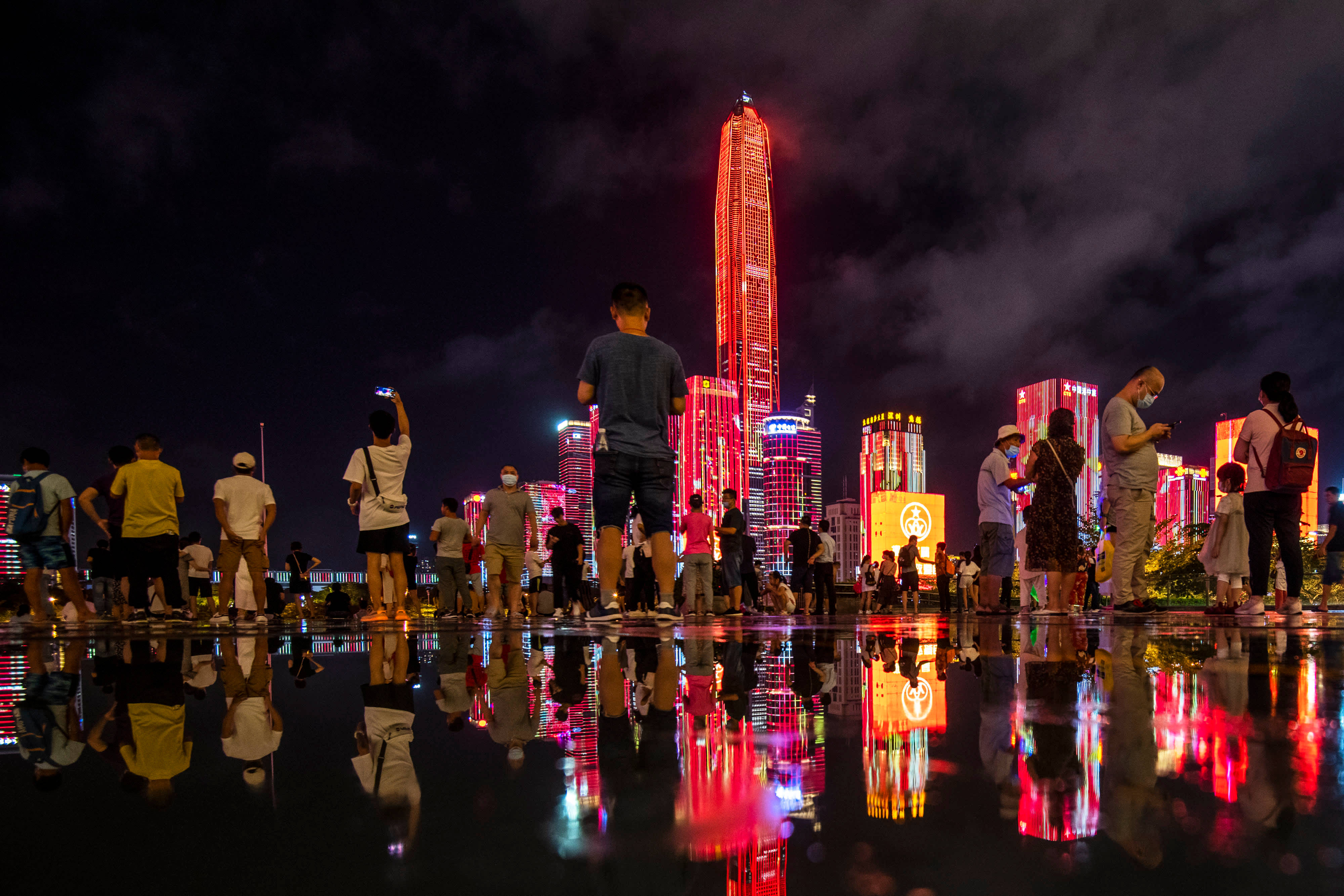 Shenzhen 桌布 壁紙 — 夜晚的人群欣賞著一座高聳的摩天大樓，發出紅光，充滿活力的城市燈光反射在潮濕的人行道上。