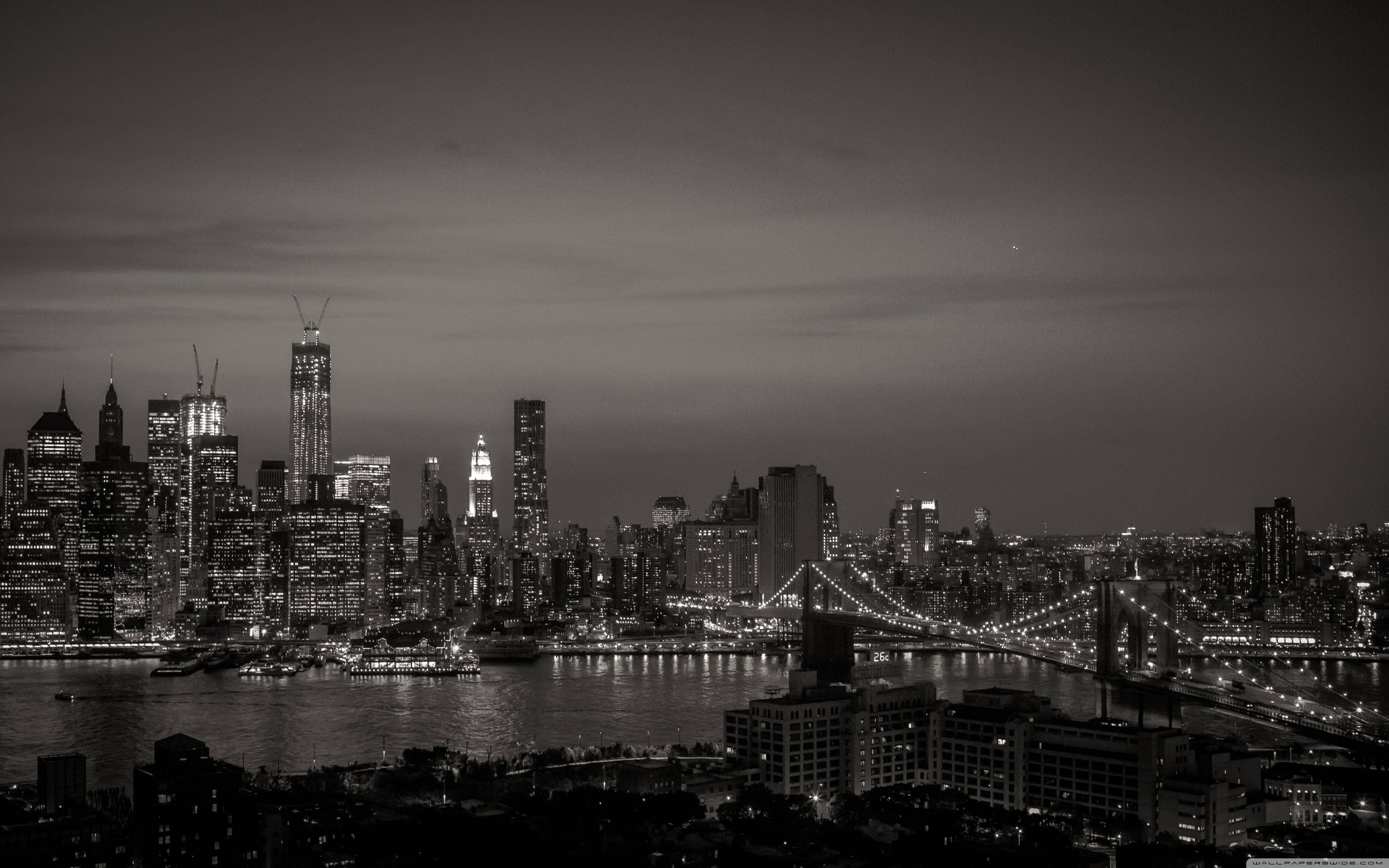 New York 桌布 壁紙 — 夜晚的單色城市景觀，有燈火通明的摩天大樓和一座橫跨黑暗河流的亮燈橋樑。