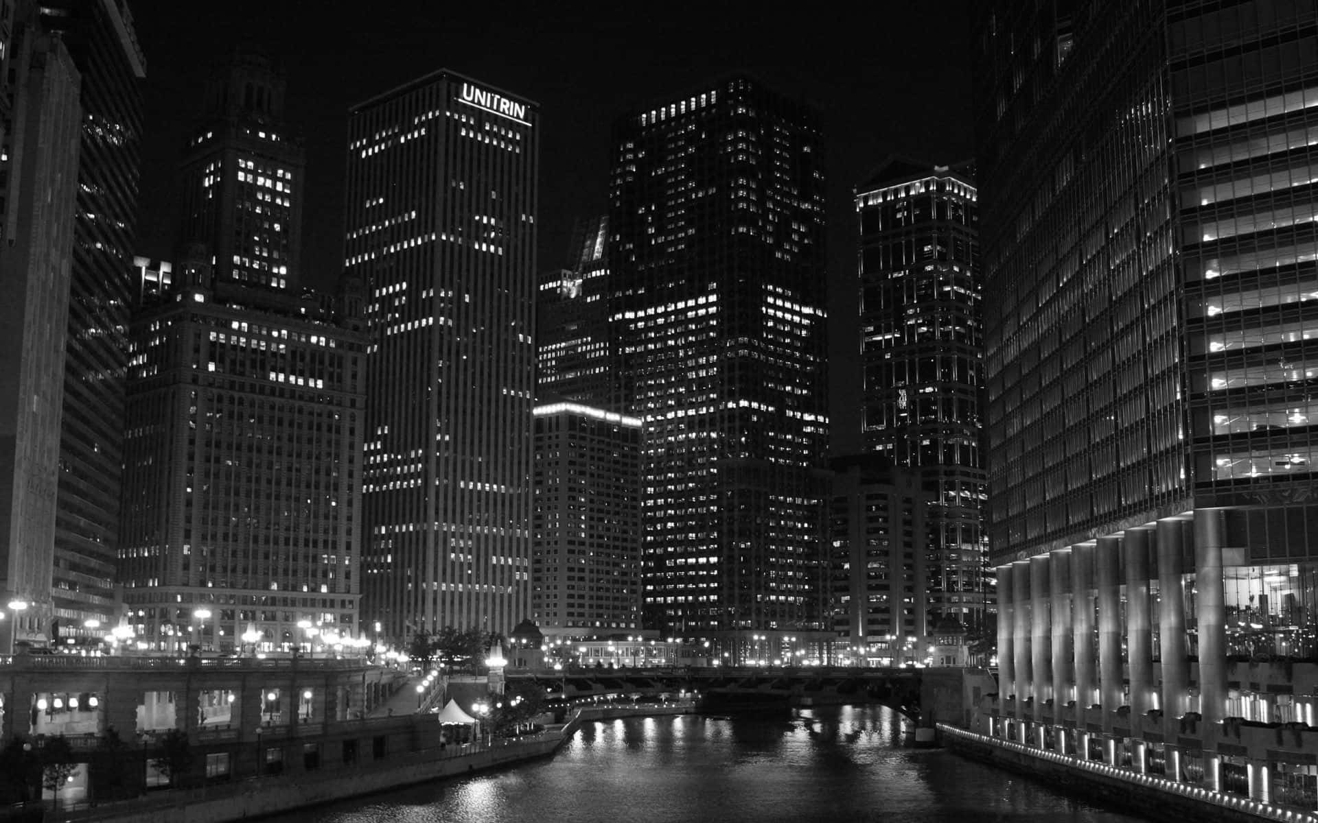 Black And White Architecture 桌布 壁紙 — 夜晚的單色城市景觀，高聳的摩天大樓將燈光反射在下面的河流上。